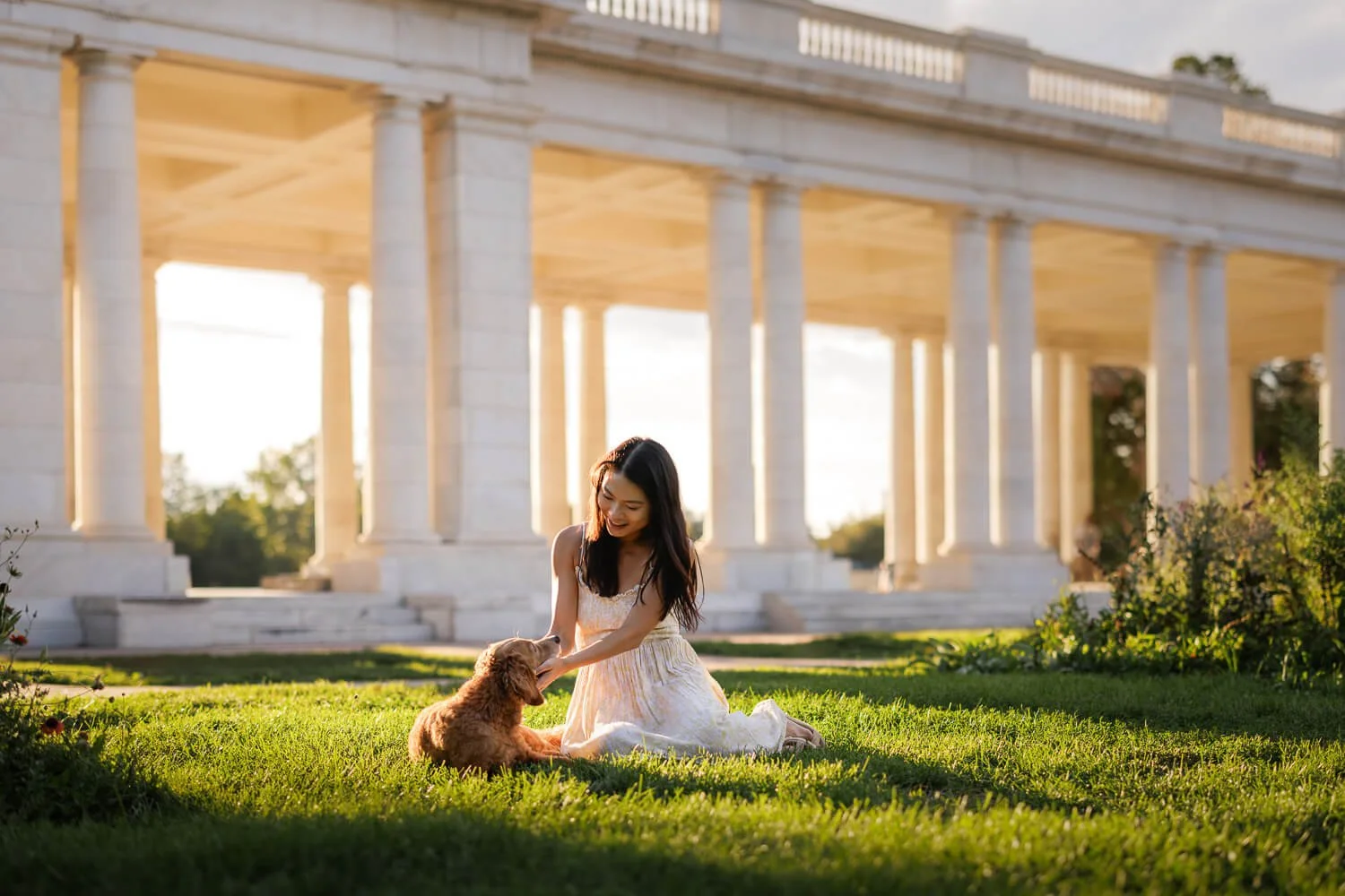 Woman in a white dress sitting on the grass petting her golden-colored dog, with the marble Cheesman Park Pavilion columns bathed in golden hour light behind them.