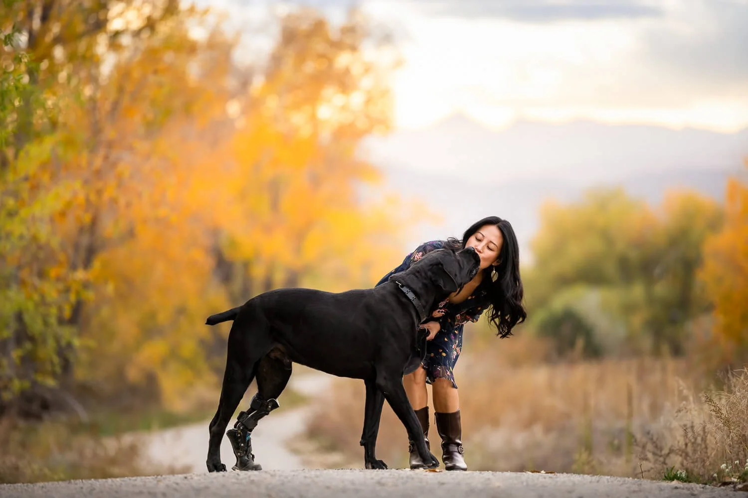 A candid moment of a woman kissing her black dog during an autumn photography session, showcasing the deep bond and connection between pets and their owners.