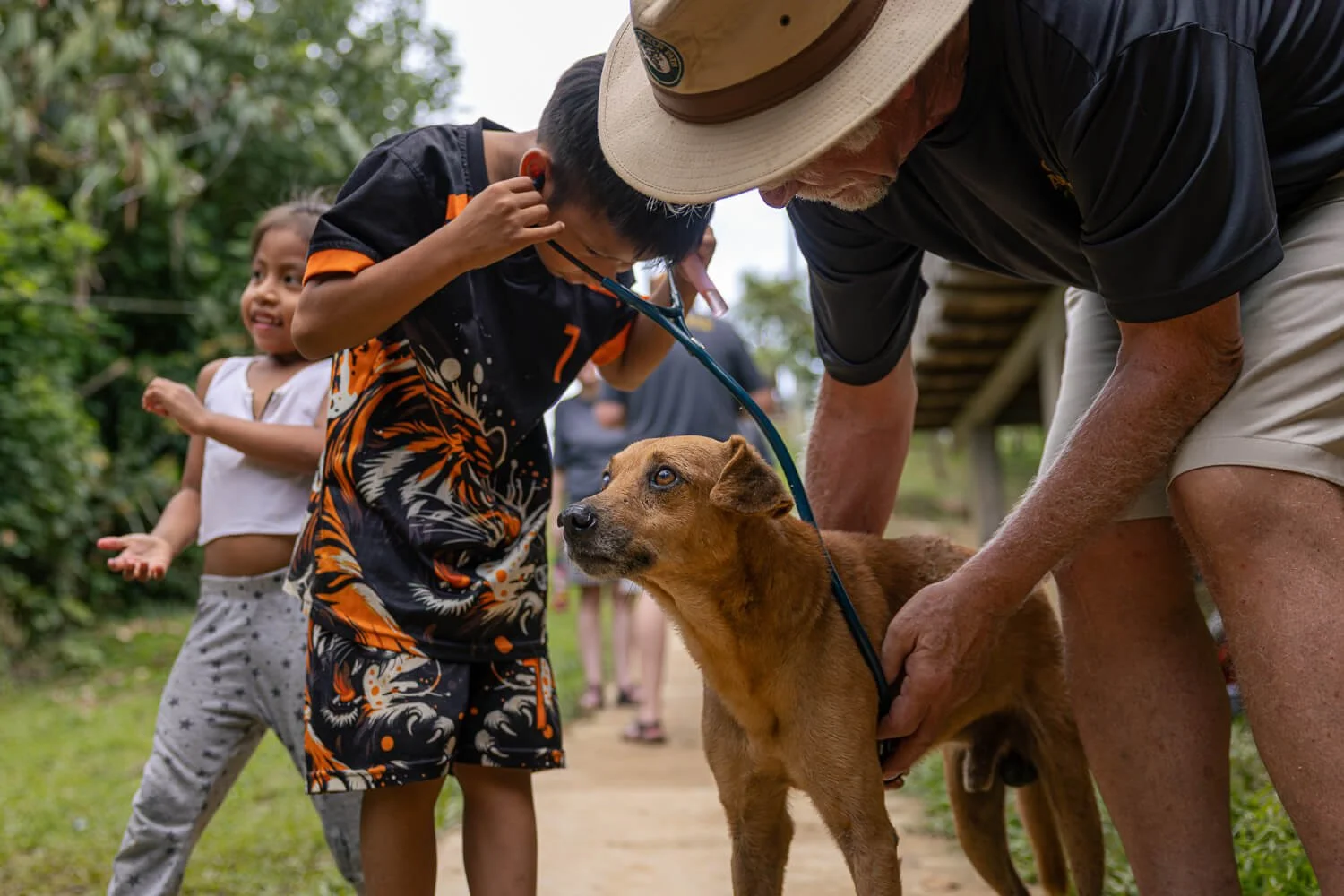 A veterinarian examines a tan dog as local children gather close to watch and help during a community vet visit in the Colombian Amazon.