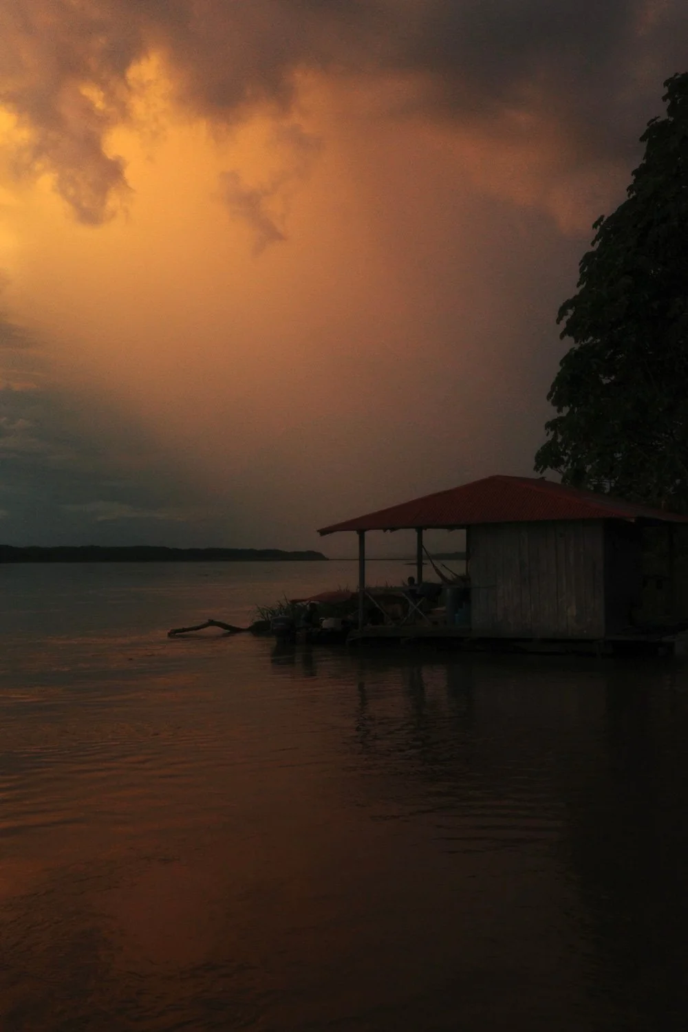 Dark storm clouds lit orange at dusk over a red-roofed stilt house on the Amazon River.