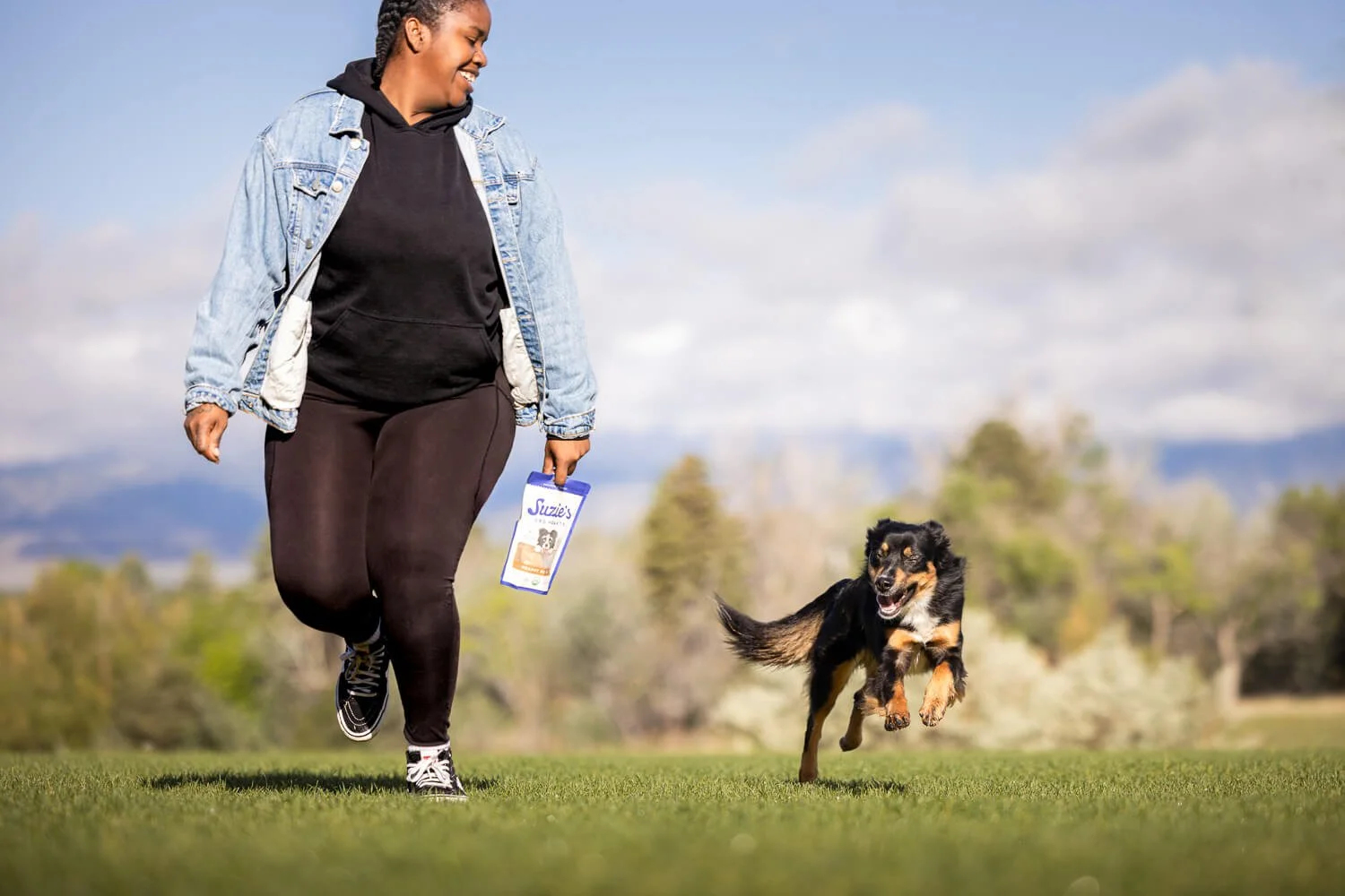 A woman in black athletic clothing and a denim jacket running on grass with a black and tan dog, possibly a Border Collie, playing fetch in an outdoor park with trees and mountains in the background.