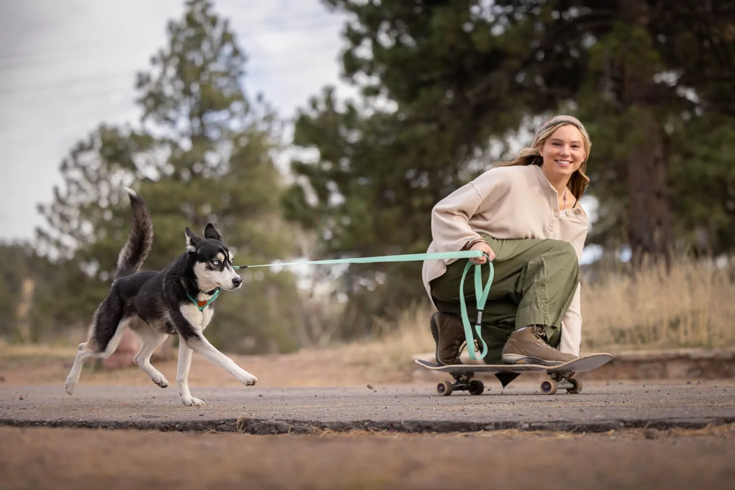 A woman riding a skateboard on an outdoor paved road while being pulled by a black and white dog wearing a teal harness and leash.