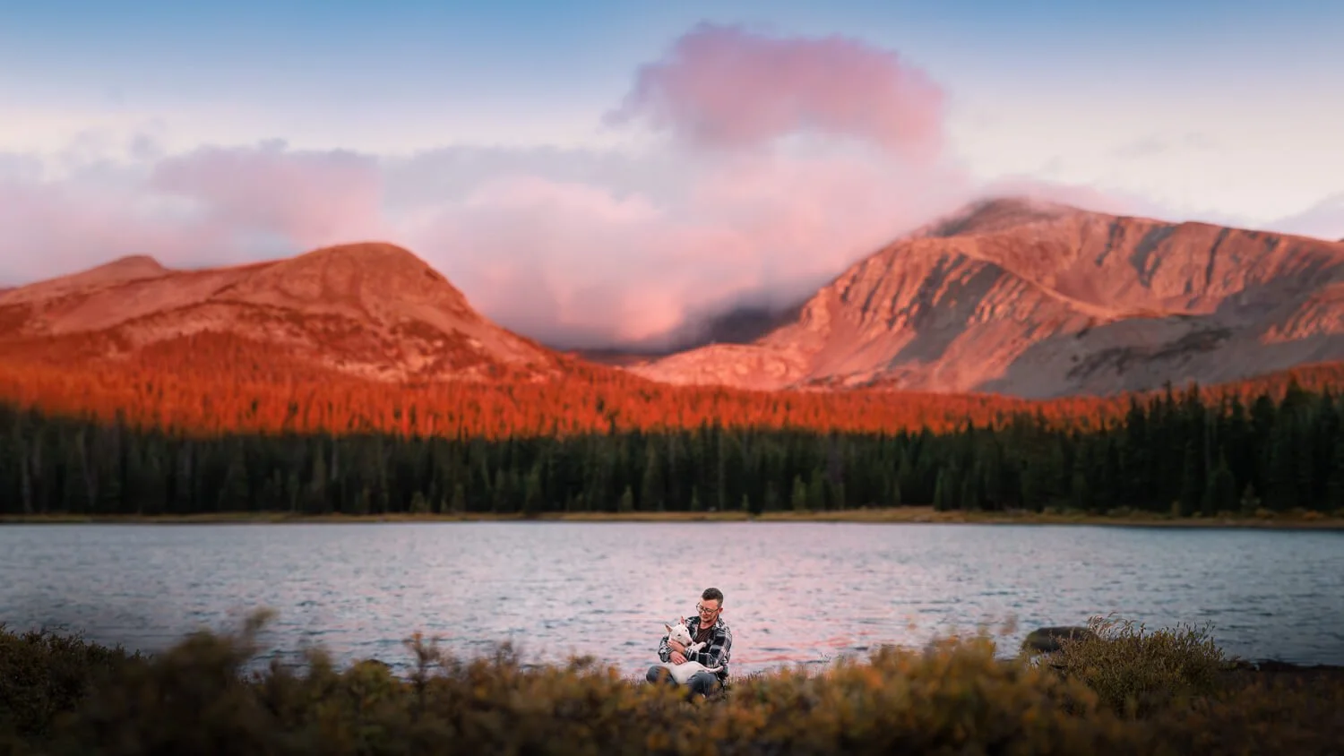 Professional pet photography of a person and dog sitting by a remote alpine lake in the Colorado Rockies during a sunset with glowing red mountain peaks.