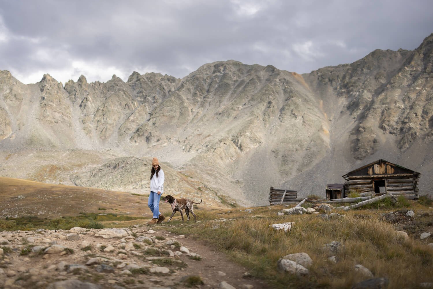 Woman and her German Shorthaired Pointer walking along a mountain trail in front of a massive rocky peak and an old rustic log cabin.