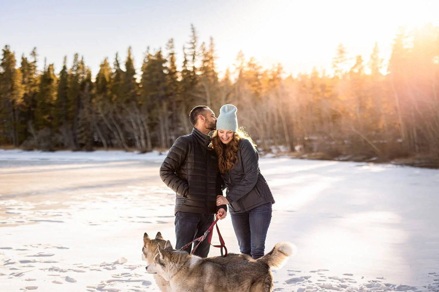 A candid and heartwarming portrait of a couple holding two Huskies on leashes in a snowy forest. The man is kissing the smiling woman on the head during a winter adventure pet photography session in Colorado.