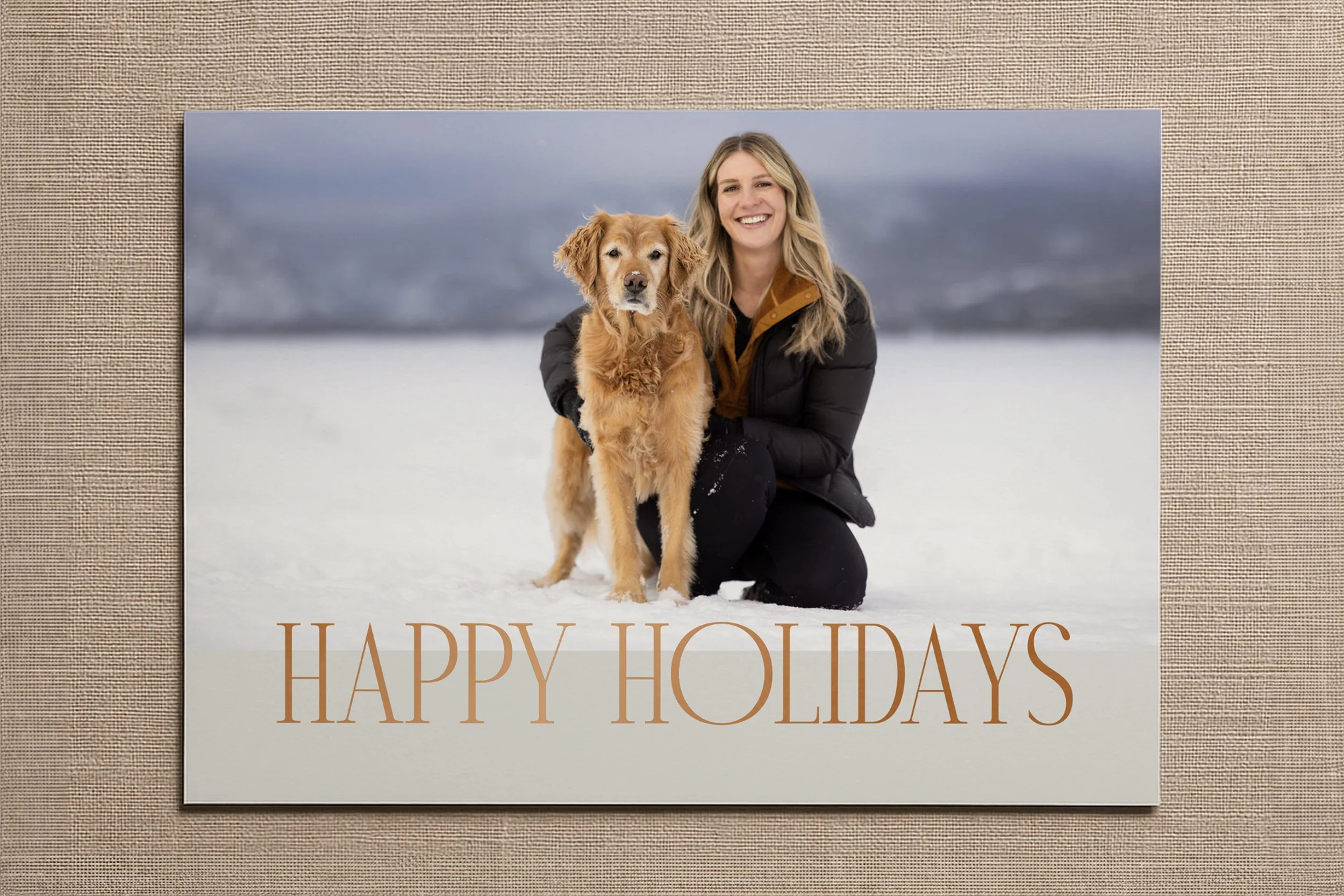 A smiling woman kneeling on snow with a golden retriever dog during winter, in front of a snowy landscape; text overlay says 'HAPPY HOLIDAYS' in brown letters.