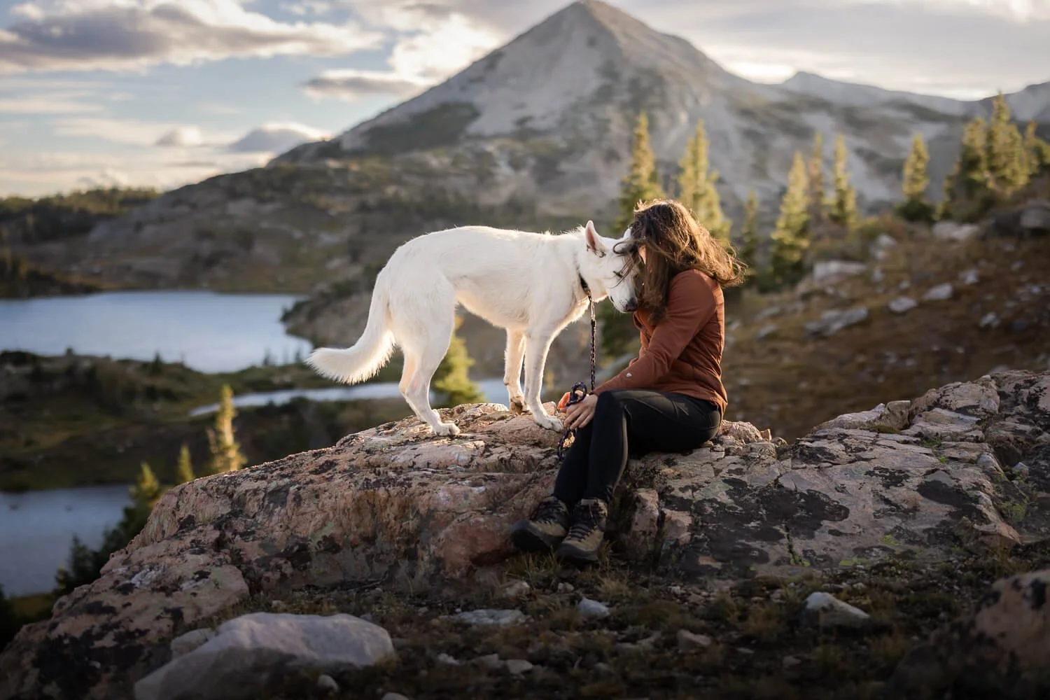 Woman sitting on a rocky mountain ridge touching foreheads with her white dog, with an alpine lake and mountain peak in the background under soft backlight.  