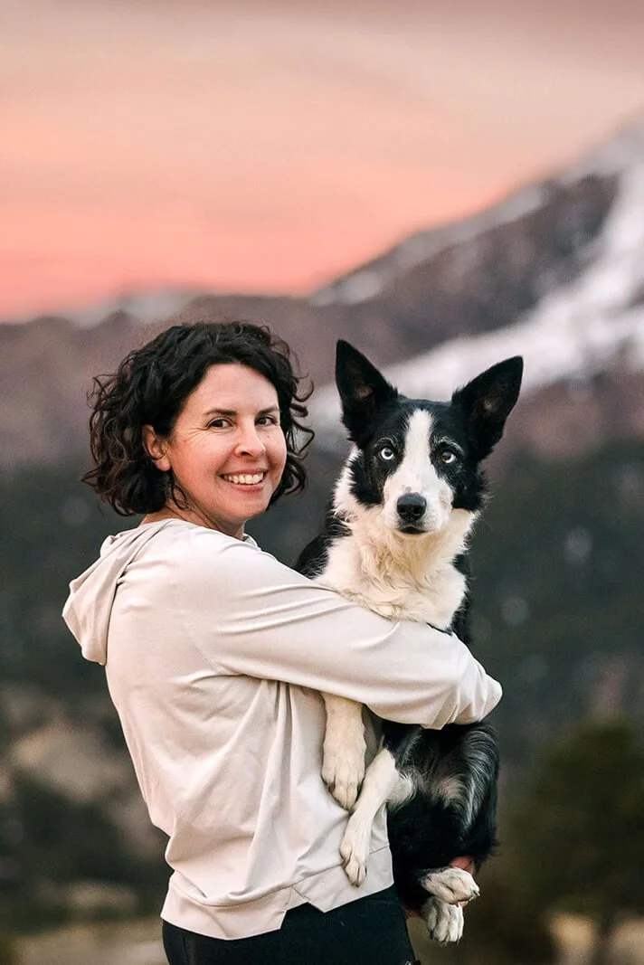 Denver pet photographer Allison Mae holding her black and white border collie against a pink sunset mountain backdrop in Colorado.