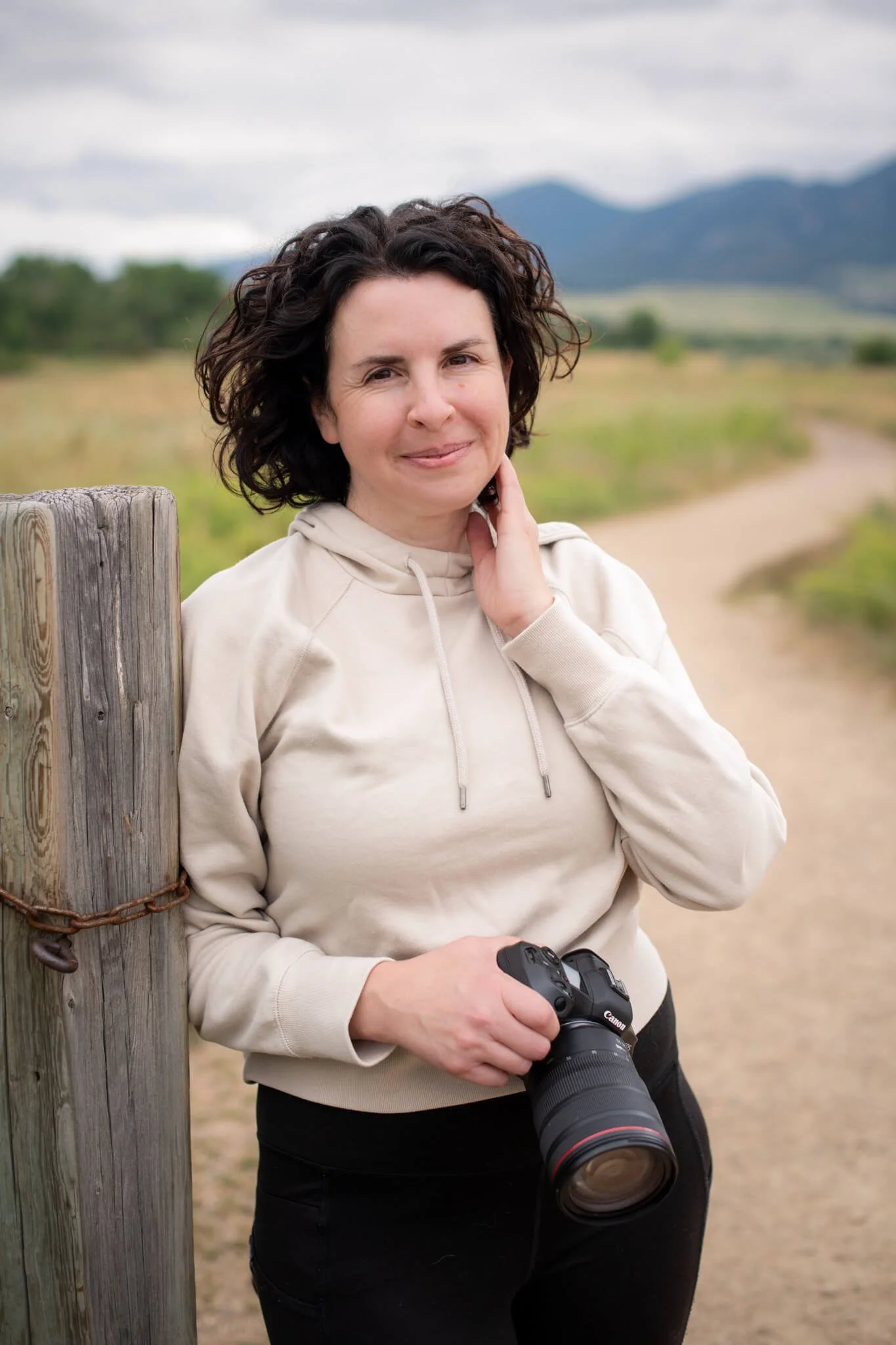 Allison, a professional Denver pet photographer, smiling and holding a Canon camera while standing on a nature trail with the Boulder mountains in the background.