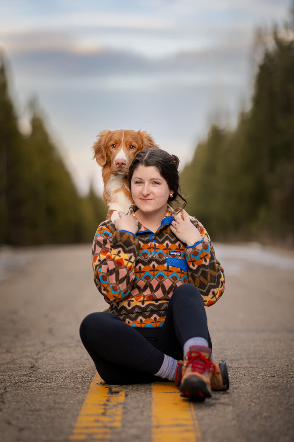 Woman sitting on the center line of a paved mountain road through a evergreen forest, holding a Nova Scotia Duck Tolling Retriever on her shoulders while both look at the camera.