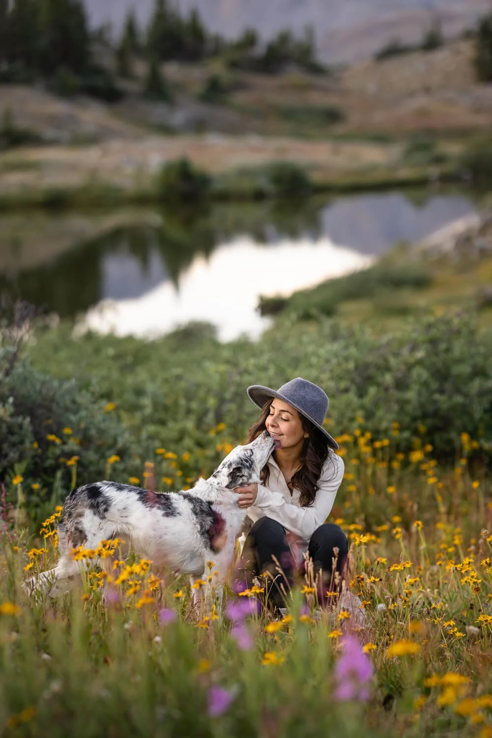 A candid moment of a dog licking its owner’s face in a field of yellow wildflowers by an alpine lake, captured by a professional Boulder pet photographer.