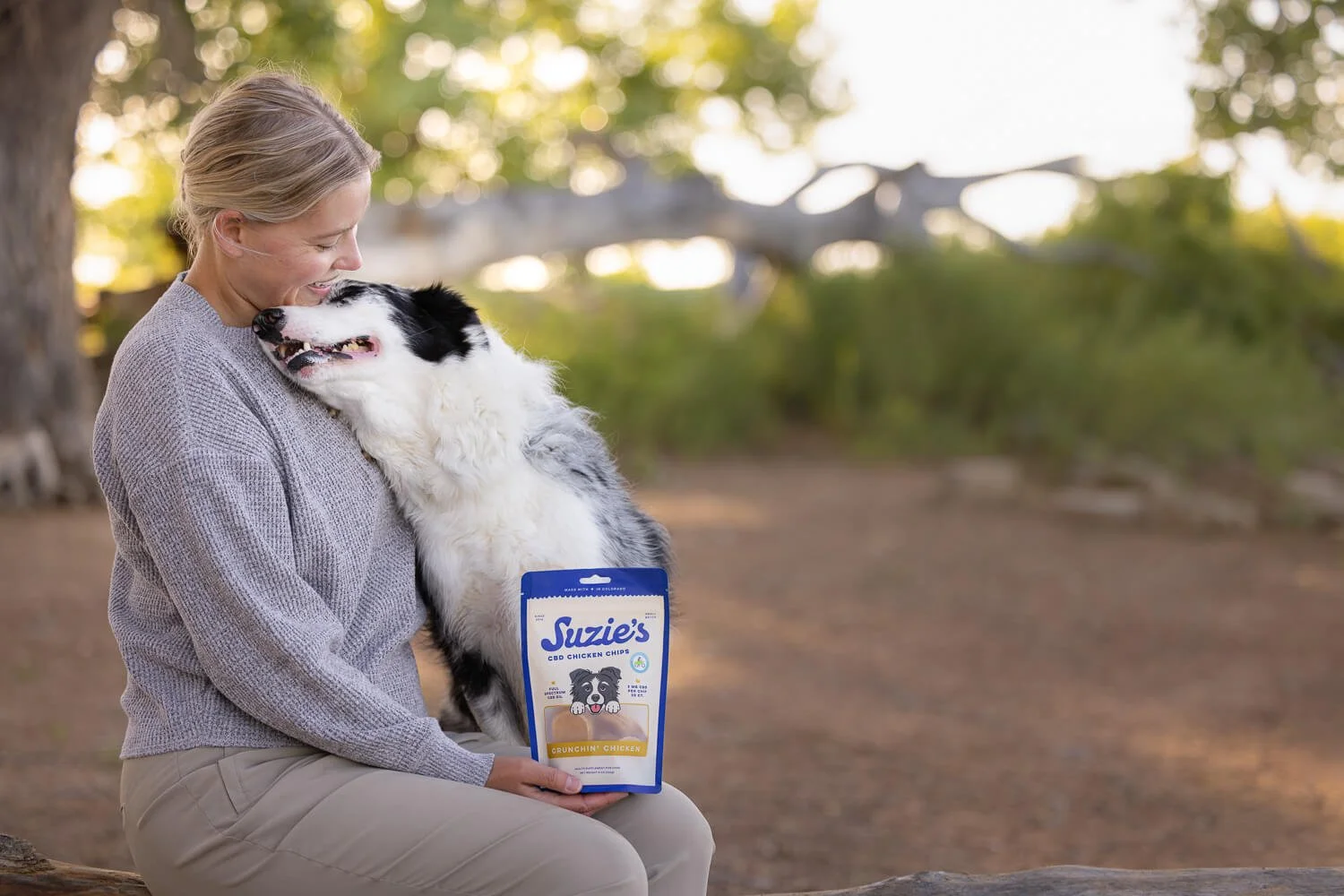A woman sitting outdoors holding a bag of Suzie's CBD chicken chips while a large black and white border collie nuzzles her face affectionately.