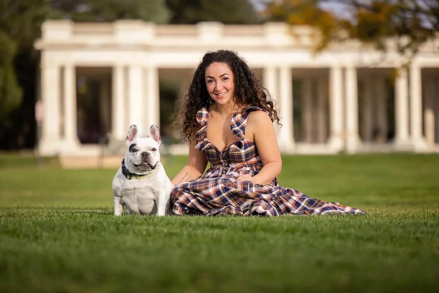 A woman and a white French Bulldog sit together on the lush green grass of Cheesman Park. In the blurred background, the historic white neoclassical Pavilion with its iconic columns stands under a bright sky.