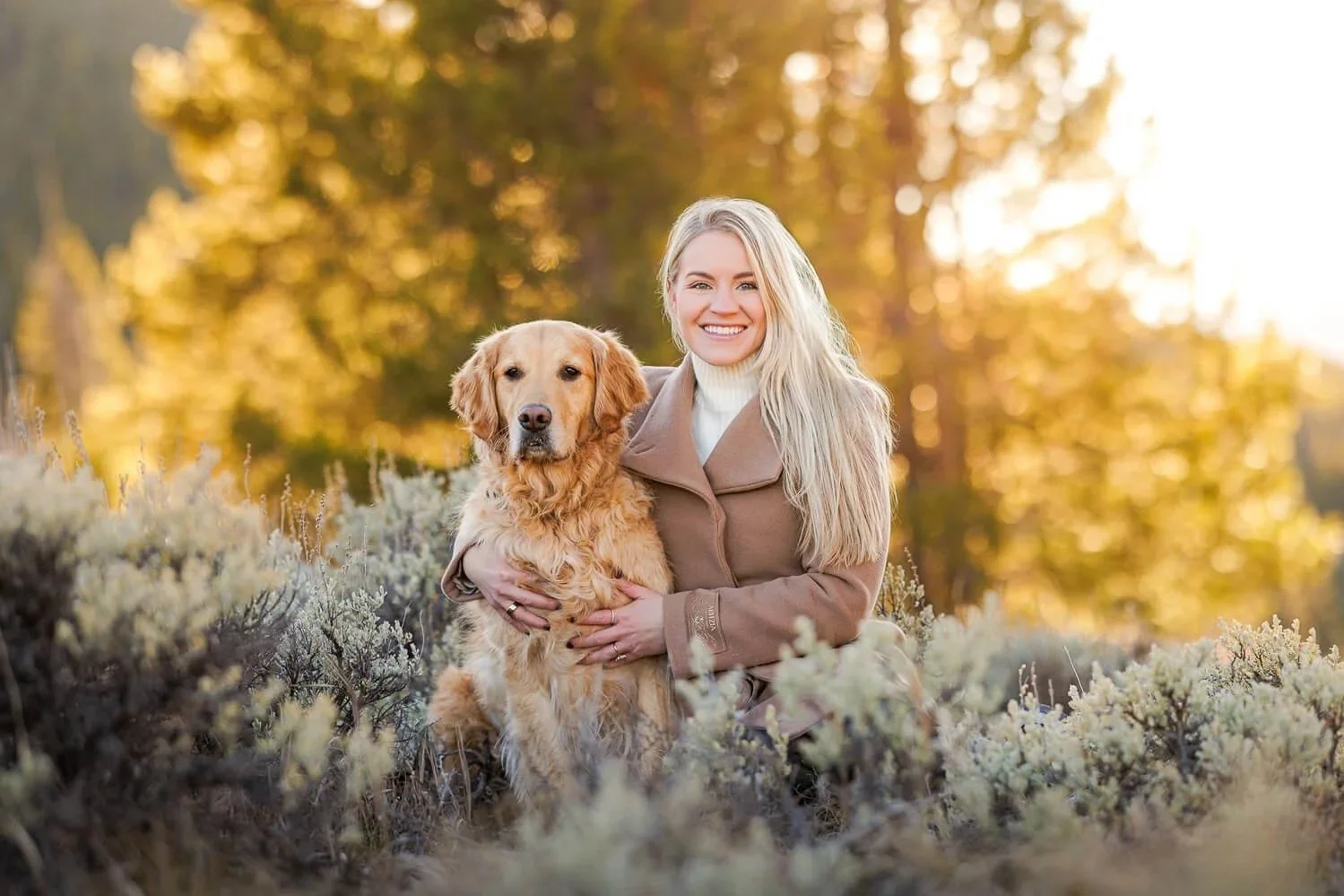 A woman in a camel wool coat and cream turtleneck posing with her golden retriever in a sun-drenched mountain landscape in Breckenridge.