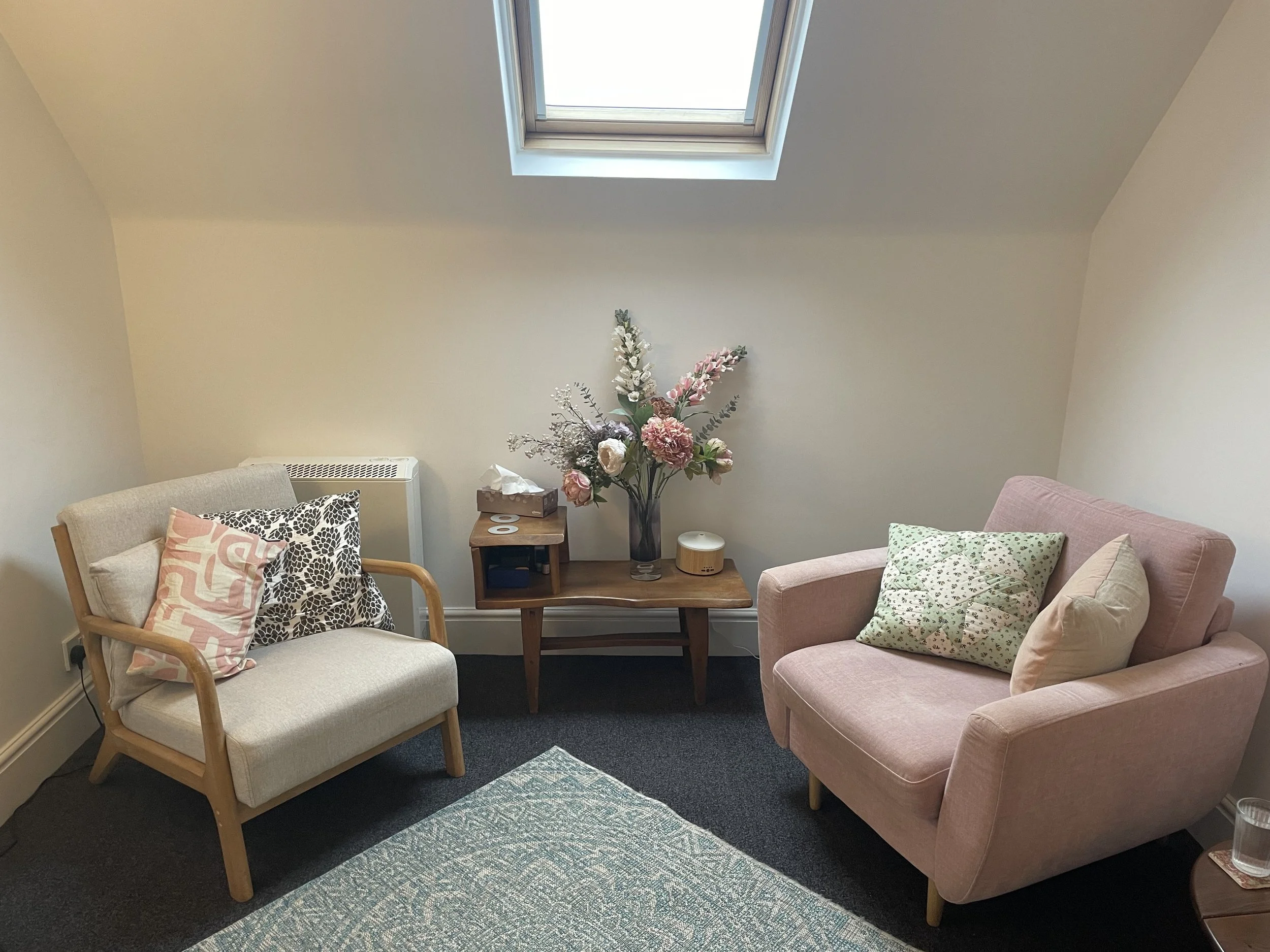 Hove therapy room with a beige armchair, pink armchair, wooden table with a vase of pink and white flowers, a tissue box, and decorative cushions.