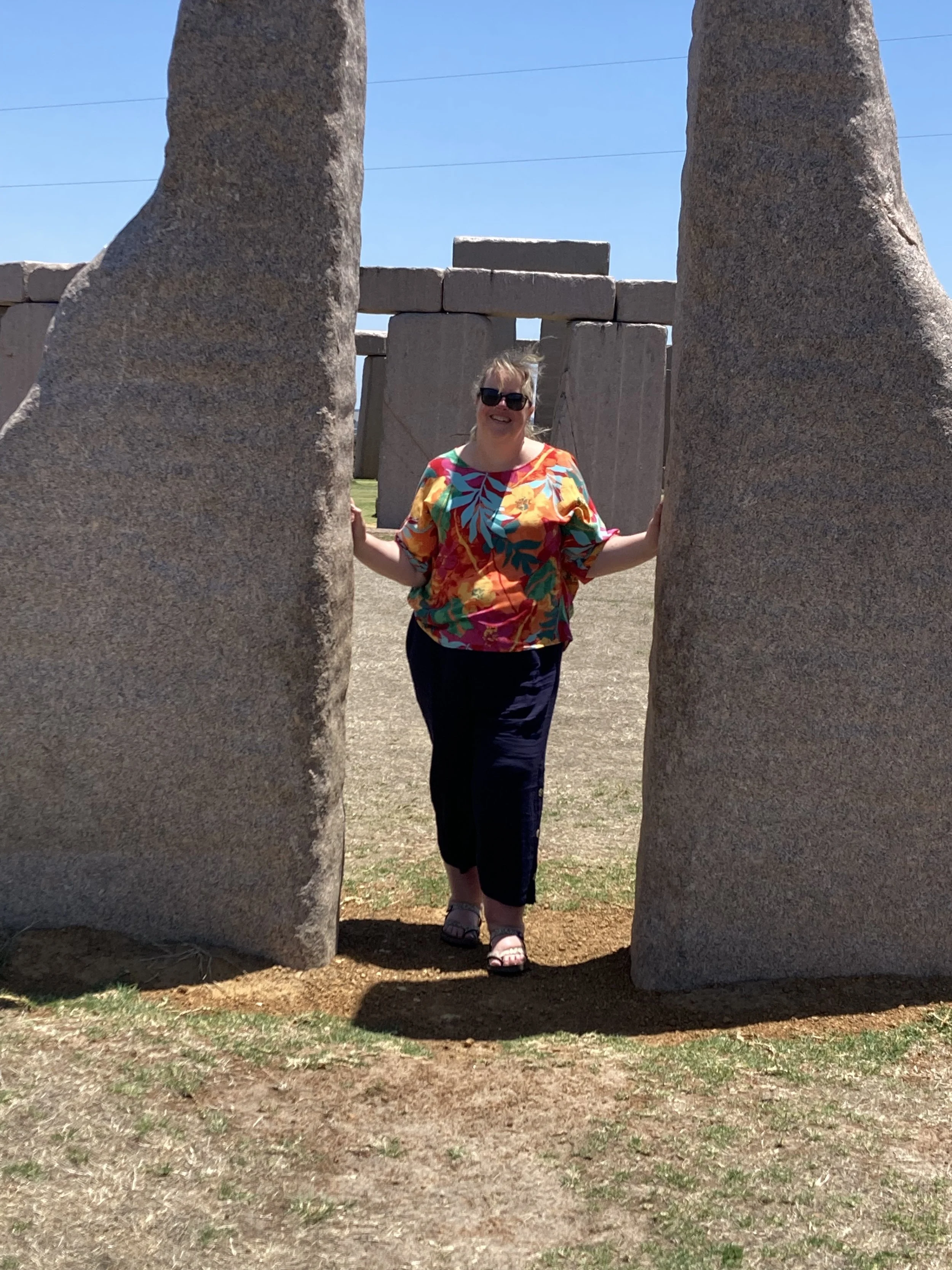 A woman standing between two large stone sculptures at Stonehenge, smiling, wearing sunglasses and a colorful floral shirt on a sunny day.