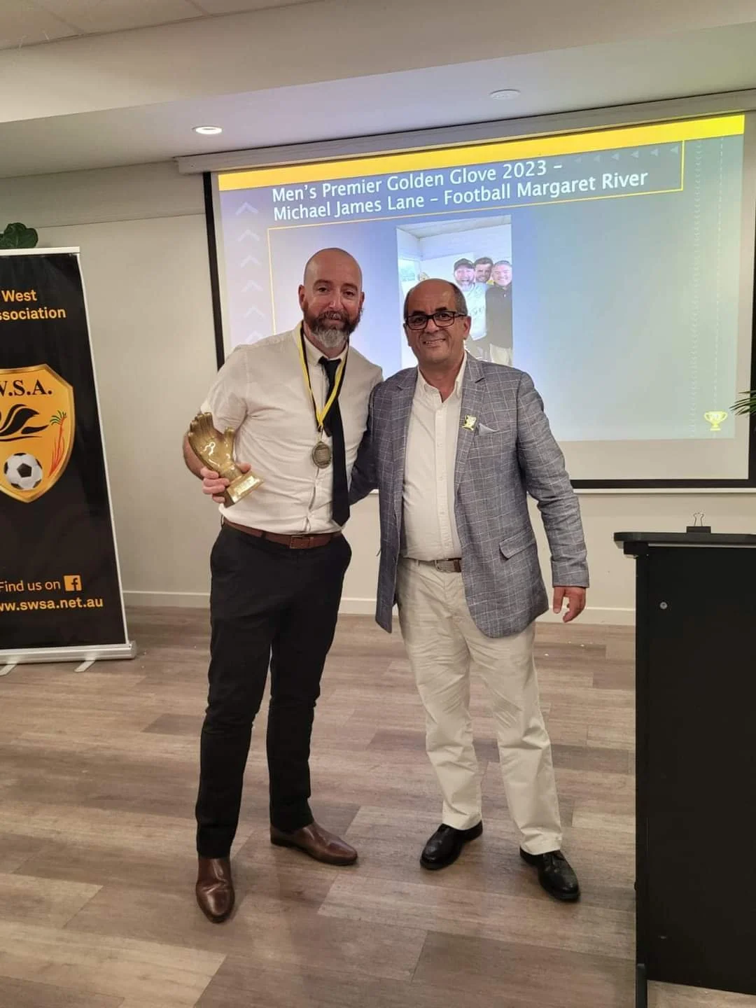 Two men standing together at an awards ceremony. The man on the left is holding a golden shoe-shaped trophy and wearing a medal around his neck. They are smiling with a presentation slide in the background that reads 'Men's Premier Golden Glove 2023 - Michael James Lane - Football Margaret River.' A banner on the left displays the logo of a soccer association.