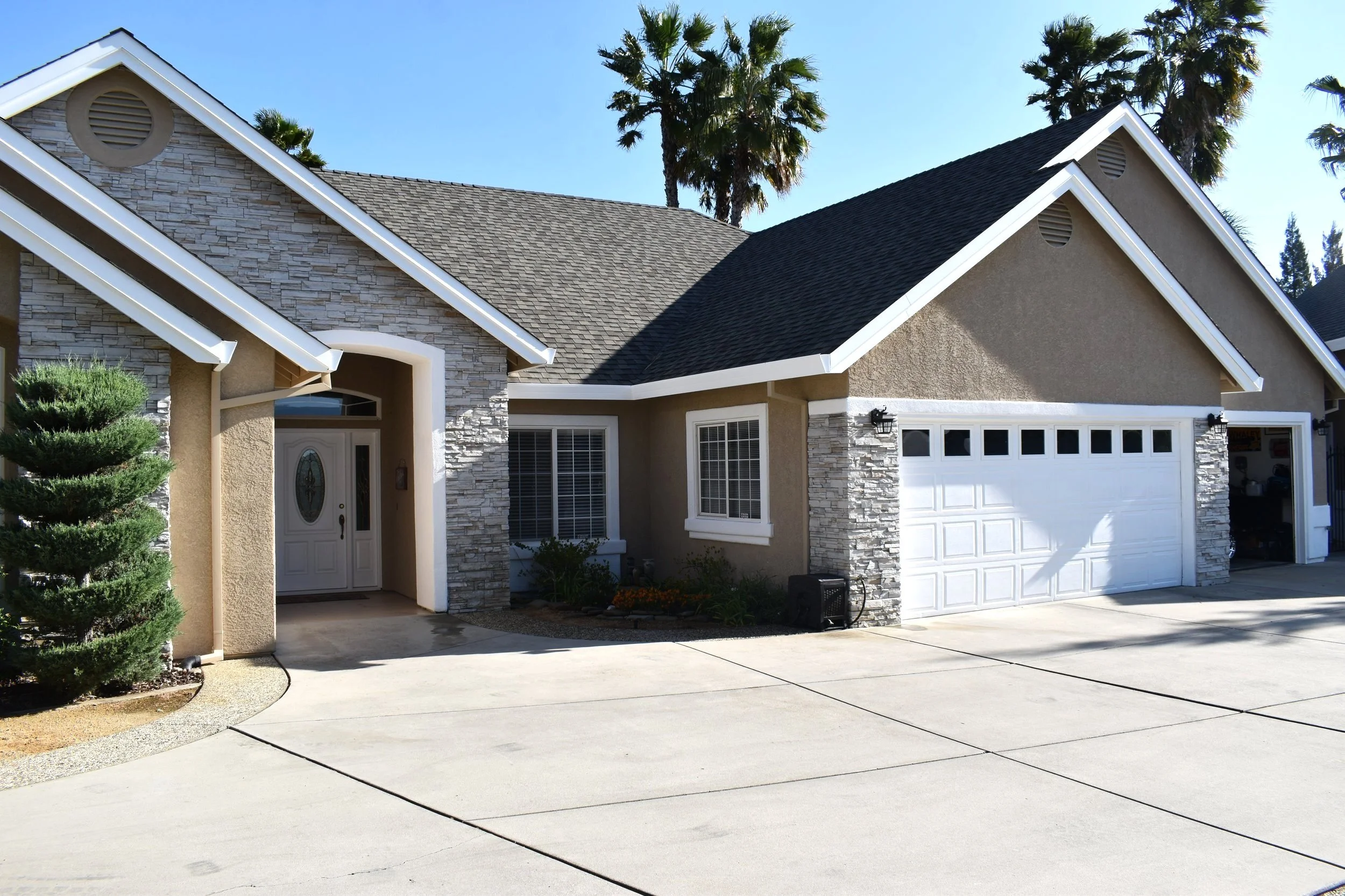 Front view of a single-family house with a concrete driveway, a white garage door, and a small landscaped garden with a sculpted tree, surrounded by trees and palm trees in the background.