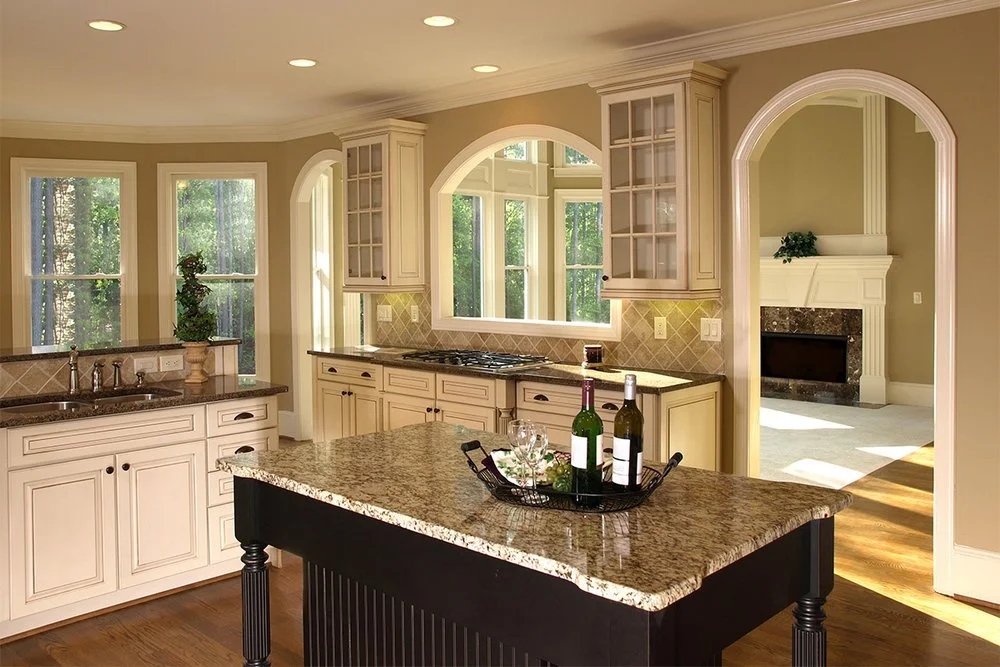 A spacious kitchen with cream-colored cabinets, granite countertops, and a central island holding wine bottles and glasses. There's an arched window above the sink, a sliding glass door, and a view into a living room with a fireplace.