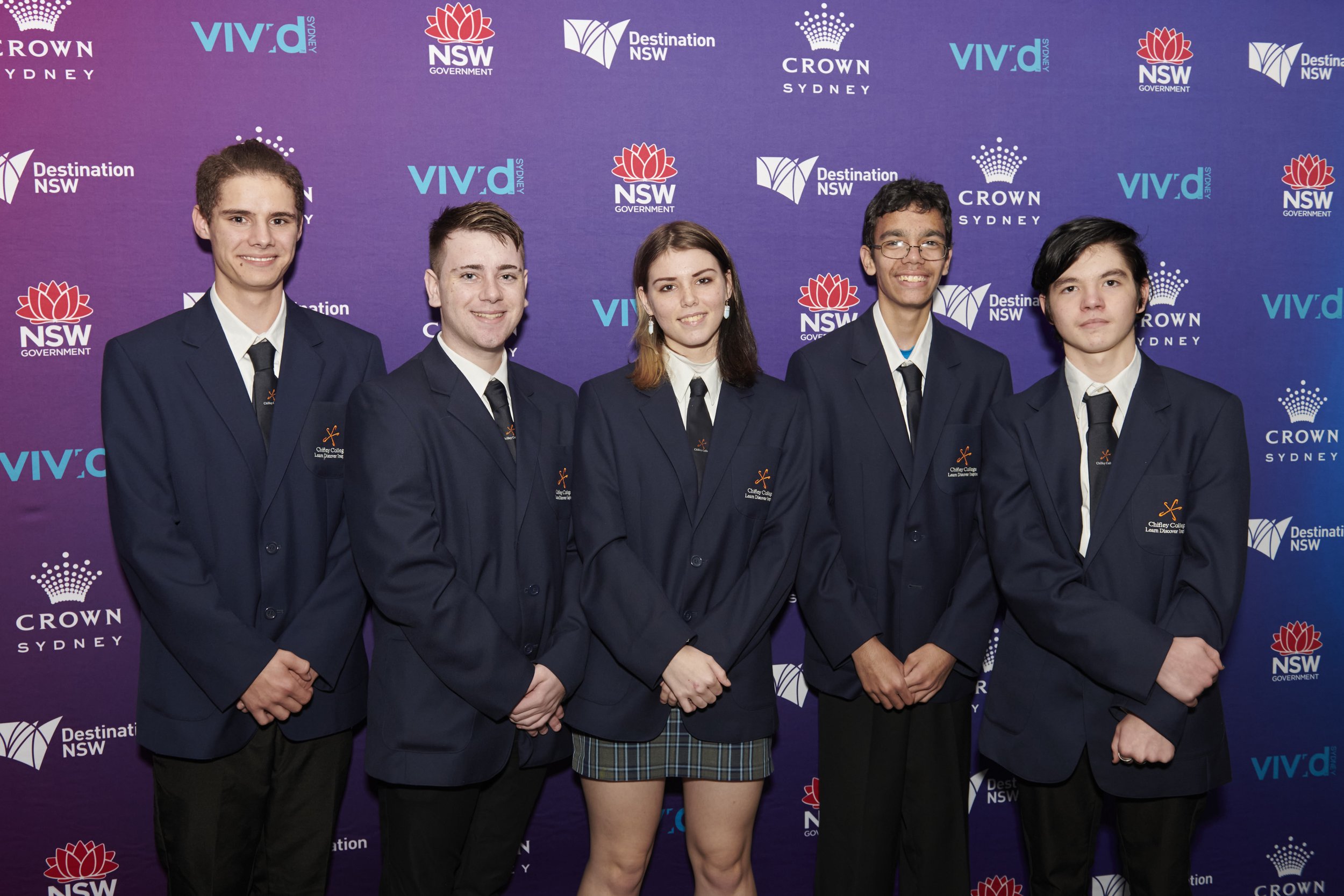 Group of five young people in formal dark blue suits and ties standing in front of a purple backdrop with logos for NSW Government, VIVID Sydney, and Crown Sydney, posing for a photo.