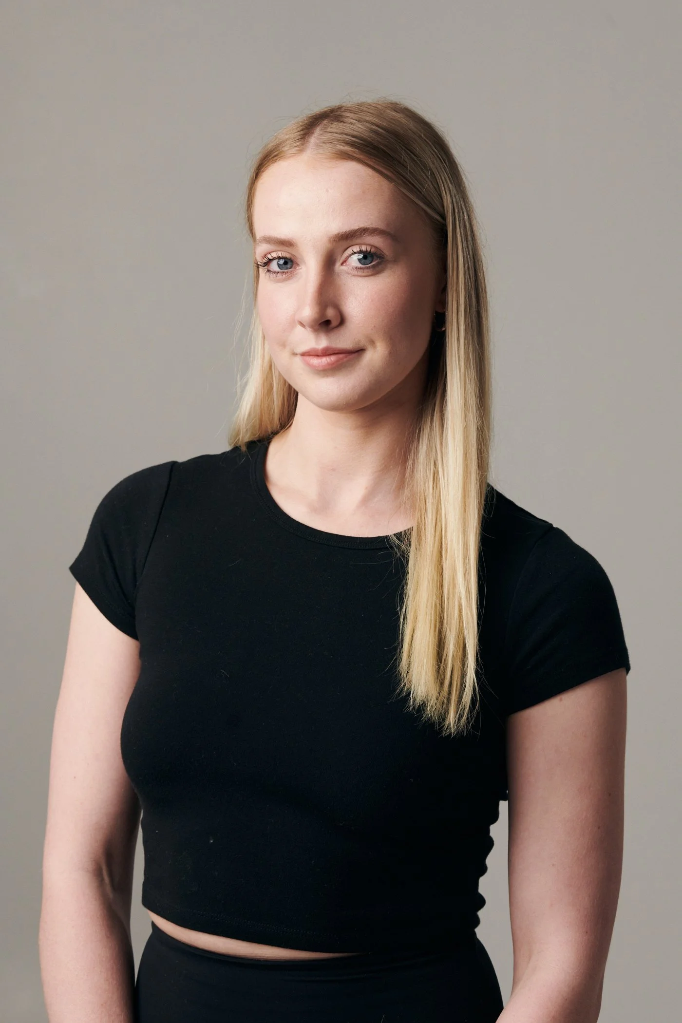 A young woman with blonde hair, blue eyes, and fair skin, wearing a black t-shirt, standing against a plain gray background.