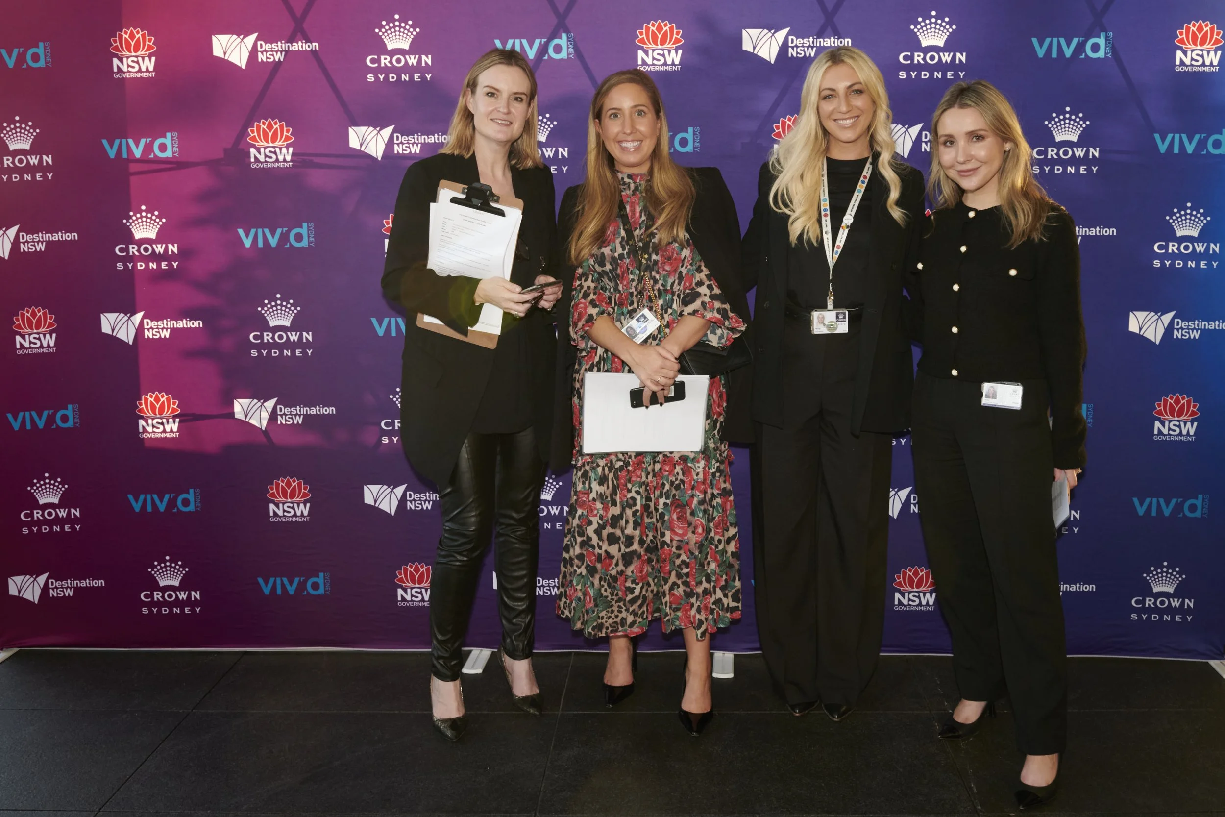 Four women standing together in front of a backdrop displaying logos of NSW Government, Crown Sydney, Destination NSW, and VIVID Sydney. They are dressed in black and colorful outfits, holding clipboards and wearing badges, at an event.