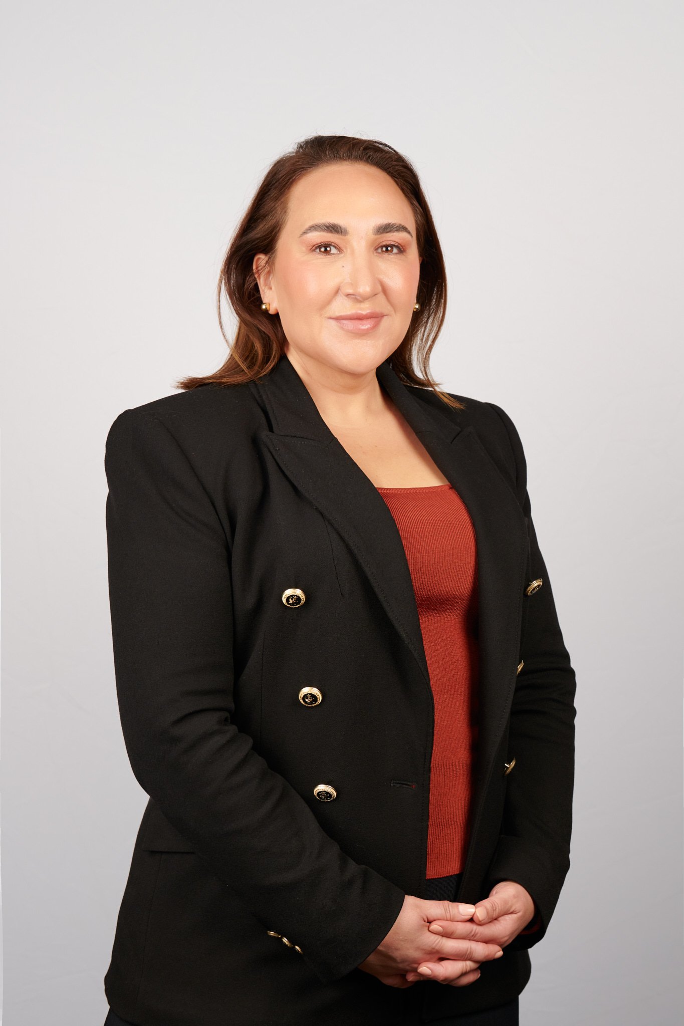 A woman with shoulder-length brown hair wearing a black blazer with gold buttons over a red top, standing with hands clasped in front against a plain light background.