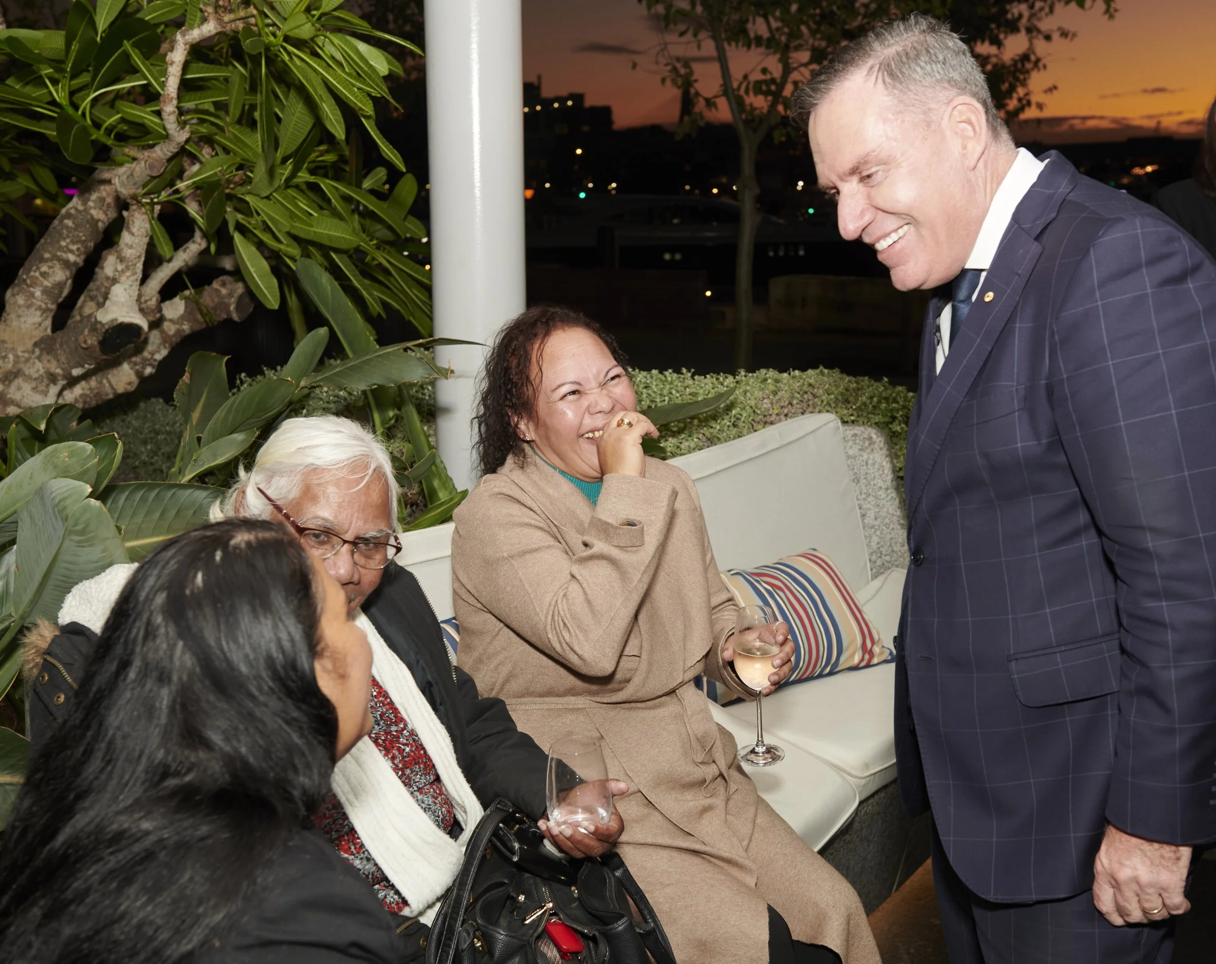 A group of people, including a man in a dark suit, smiling and engaging in conversation, at an outdoor gathering during sunset, with lush greenery and a cityscape visible in the background.