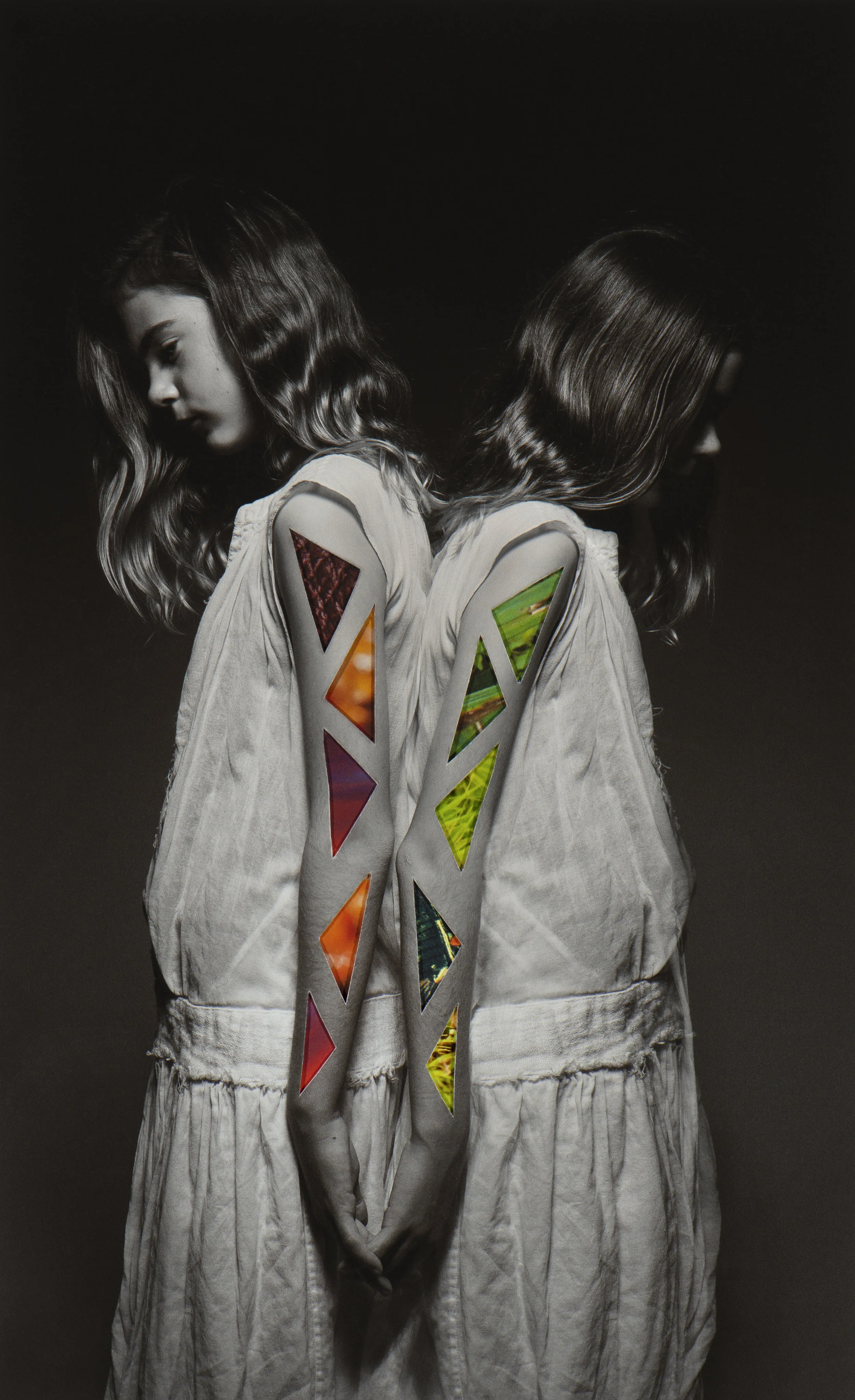 A black-and-white photo of two young women standing back to back with their heads slightly bowed. The women are wearing matching light-colored outfits with colorful, geometric-patterned cutouts on their sleeves.