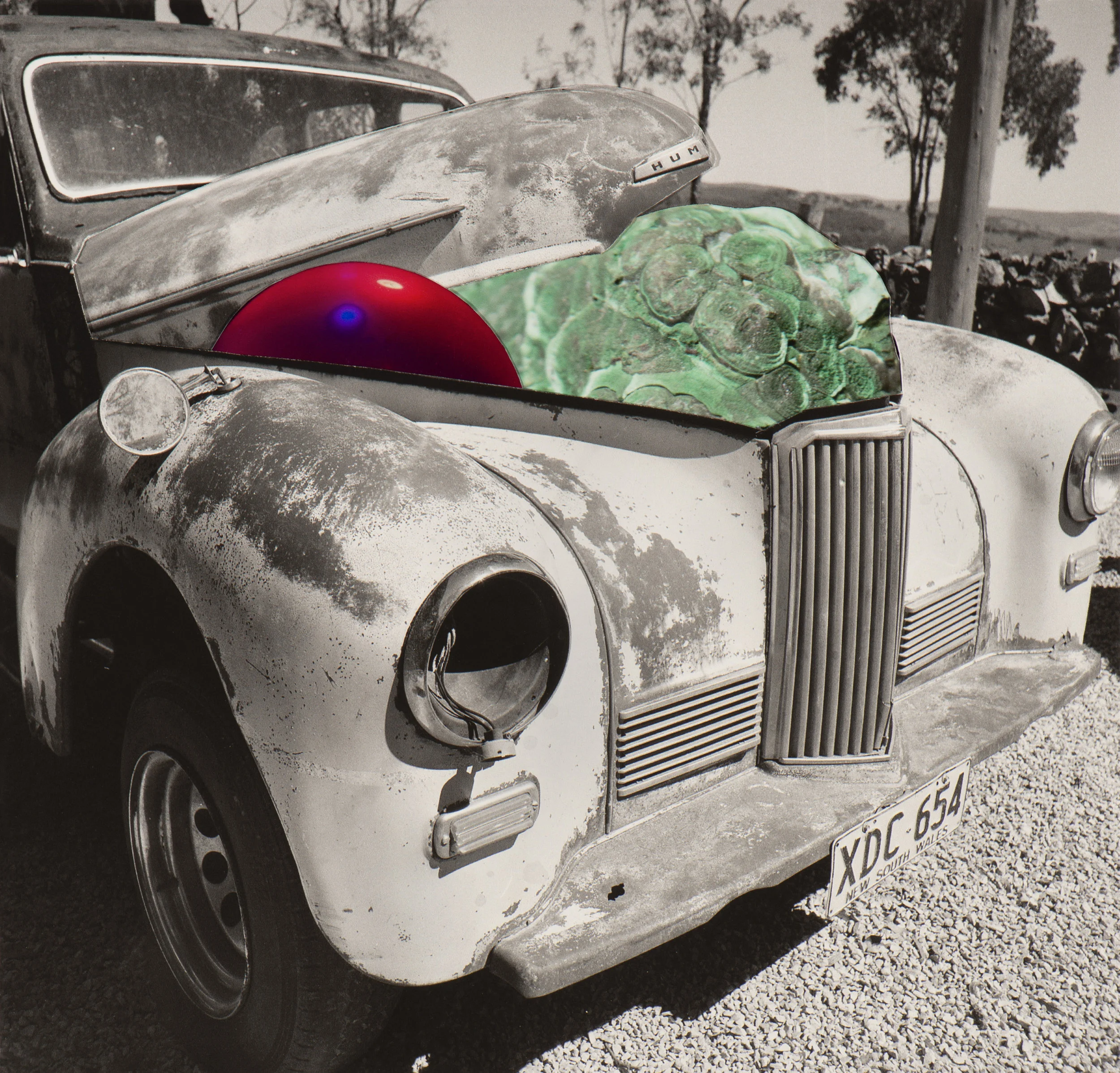 A weathered vintage car with a South Wales license plate, filled with a red and a green object, on a gravel surface outdoors with trees and a stone wall in the background.