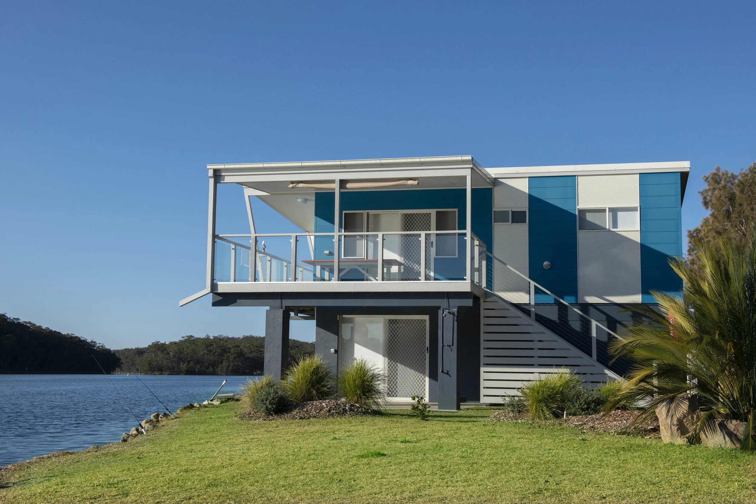 Modern two-story house with blue and white exterior, balcony, and staircase near a lake with green grass and trees.