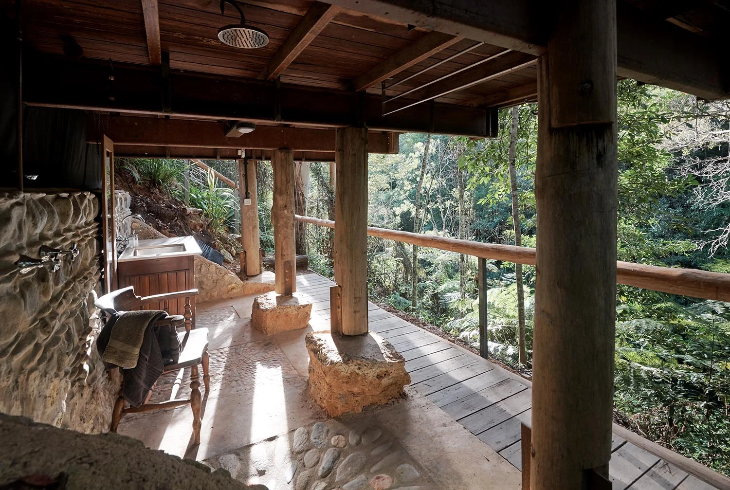 Outdoor porch area with wooden posts, a stone wall, a white chair with a towel, a sink, and a view of lush green trees and foliage outside.