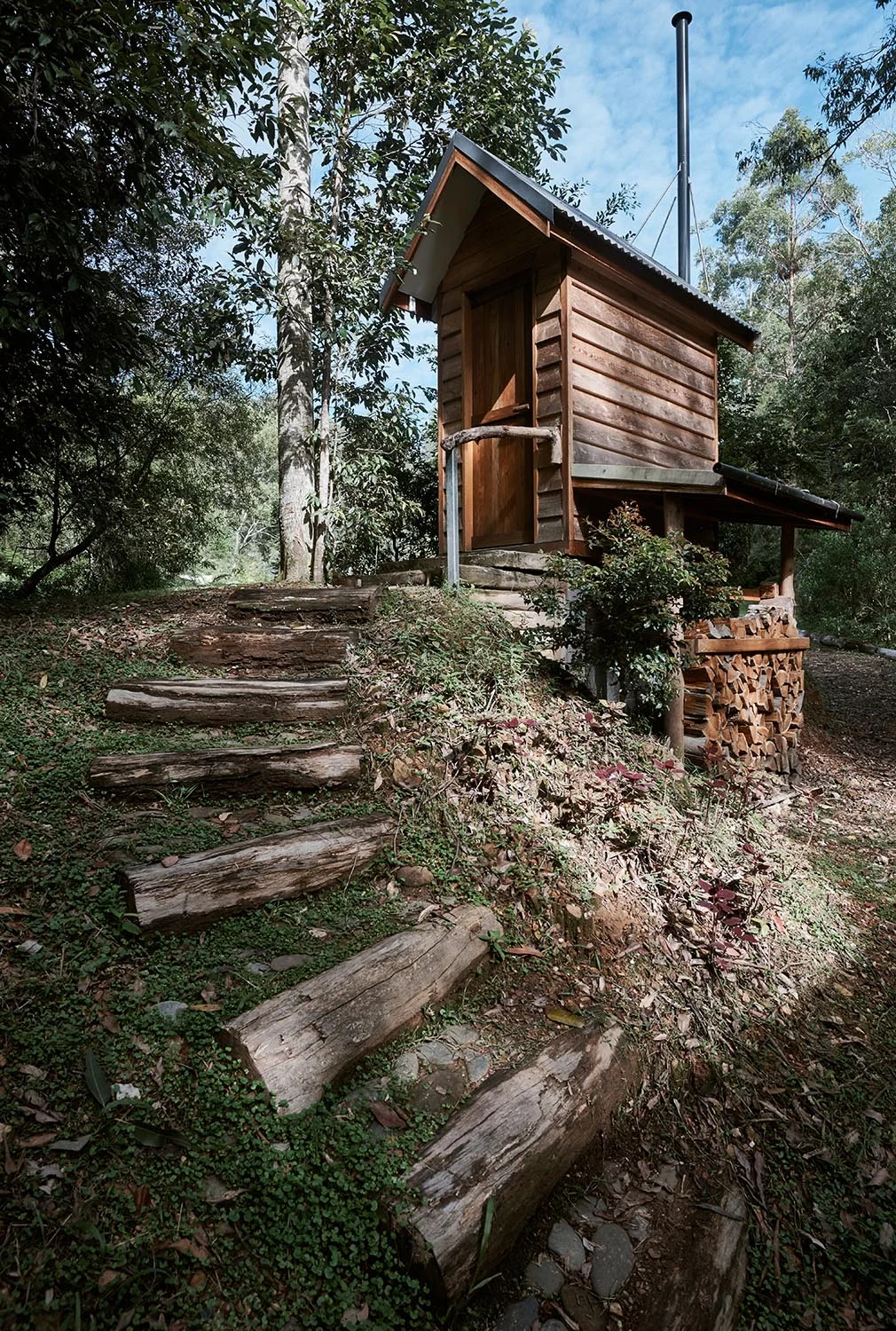Wooden treehouse built on stilts with a metal chimney pipe, surrounded by trees and forest, with wooden steps leading up to the entrance.