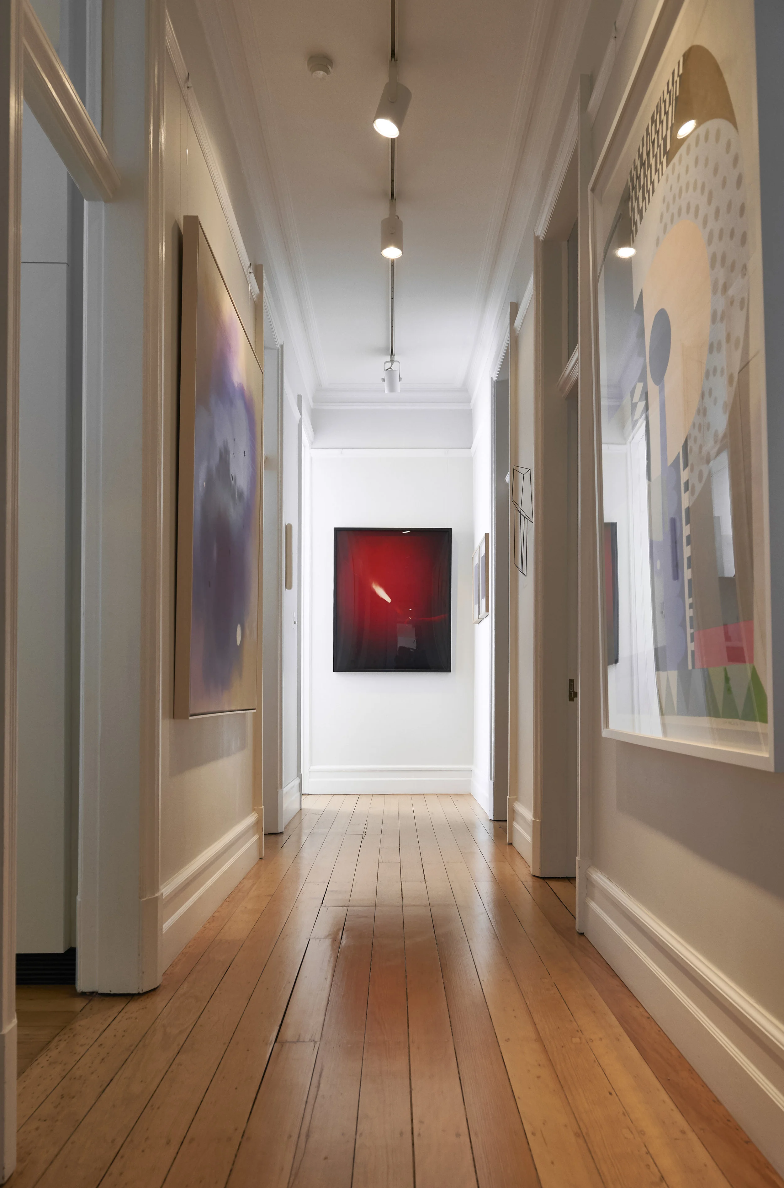 View of an art gallery hallway with framed artwork on white walls, wooden floor, and ceiling track lighting.