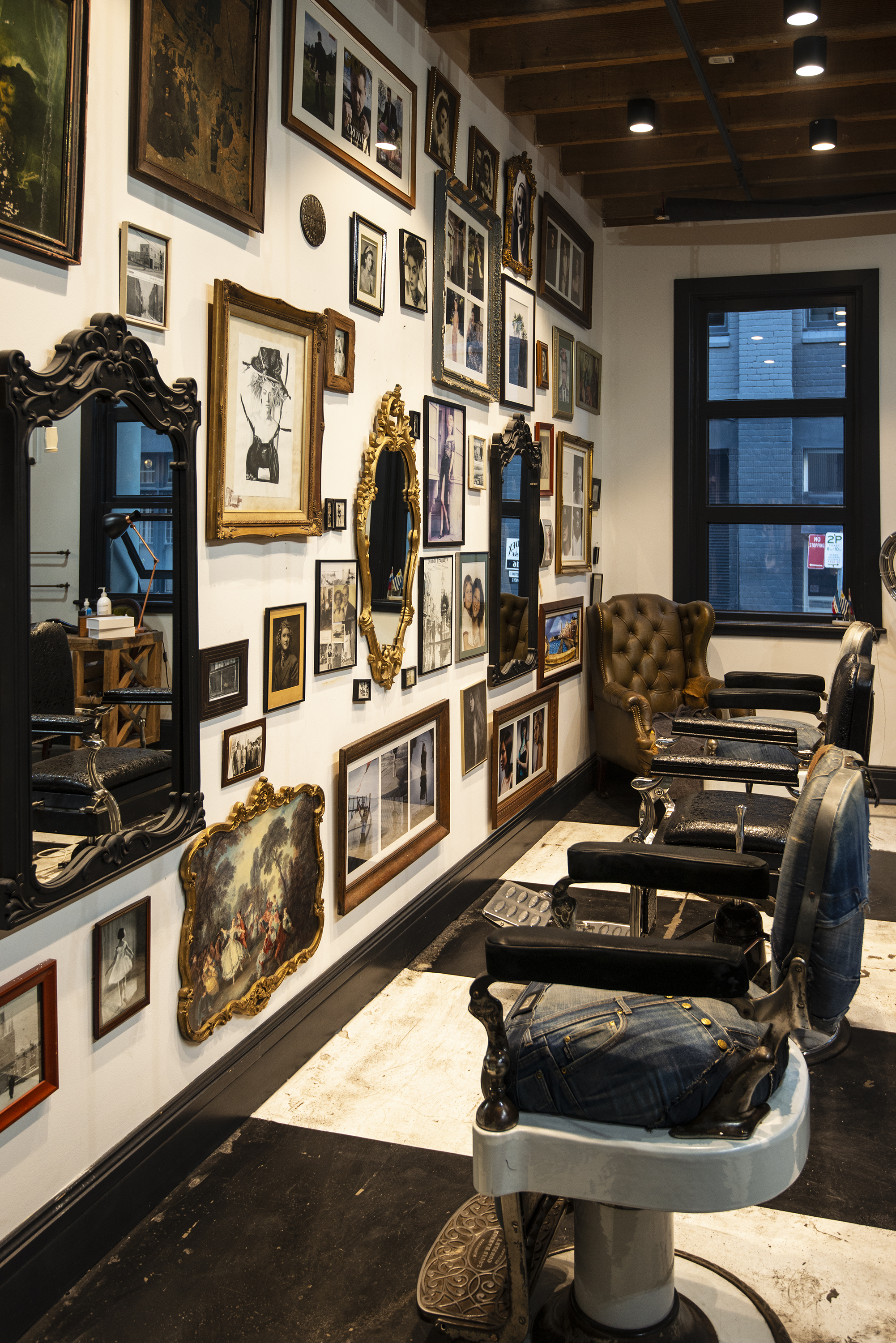 Art gallery with a white wall decorated with various framed pictures, paintings, and mirrors, black chairs, leather armchair near a window, and vintage barber chair with jeans on it.