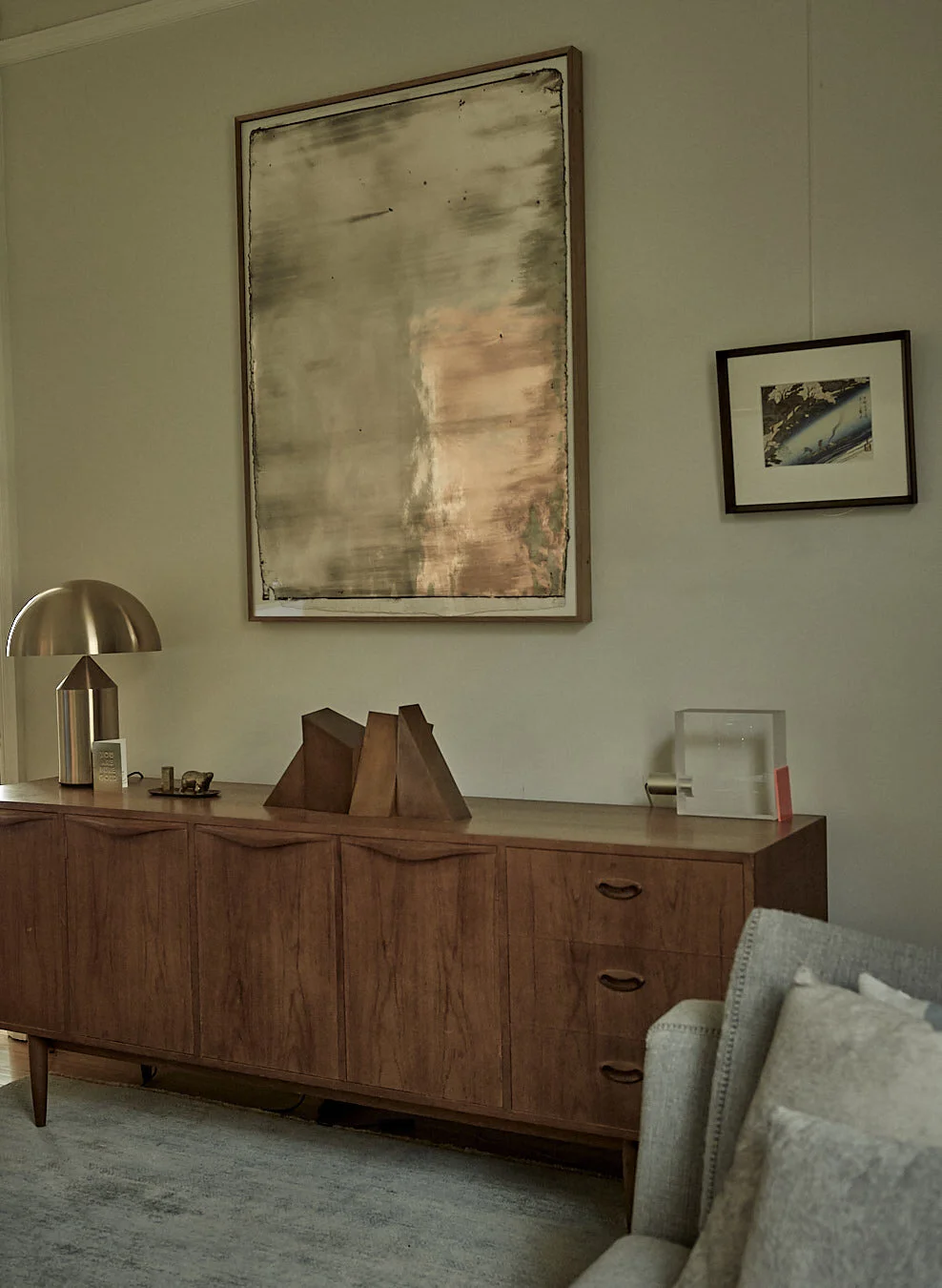 Mid-century modern wooden sideboard with decorative objects and framed artwork above, beige wall, gray sofa with pillows, area rug, gold dome-shaped table lamp.