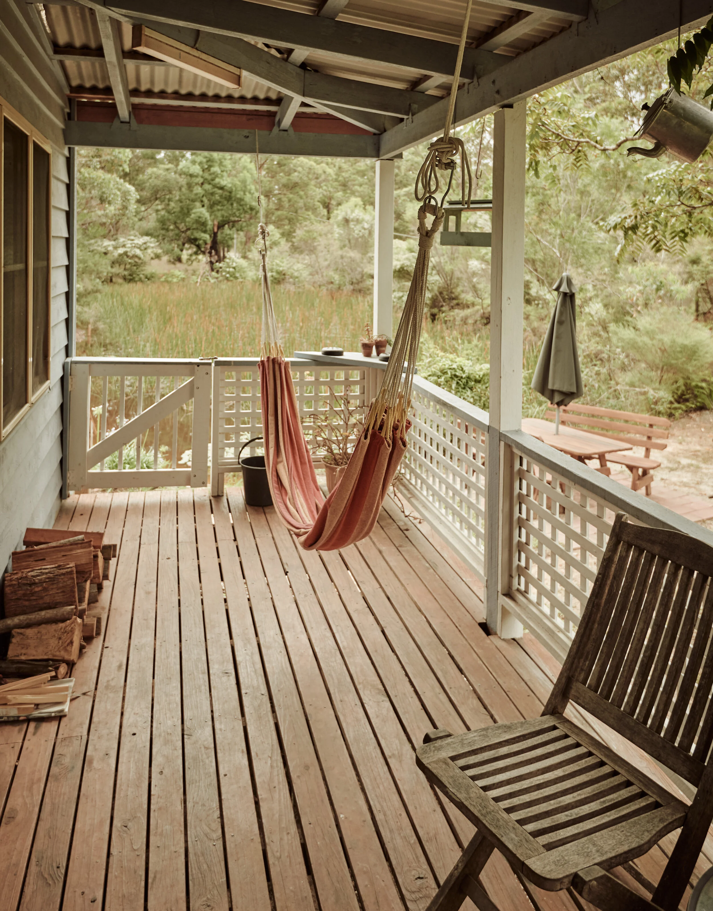 A cozy wooden porch with hanging pink and beige hammocks, a wooden chair, a small table with potted plants, a stack of firewood, and a grill with a closed umbrella outdoors in a lush green area.