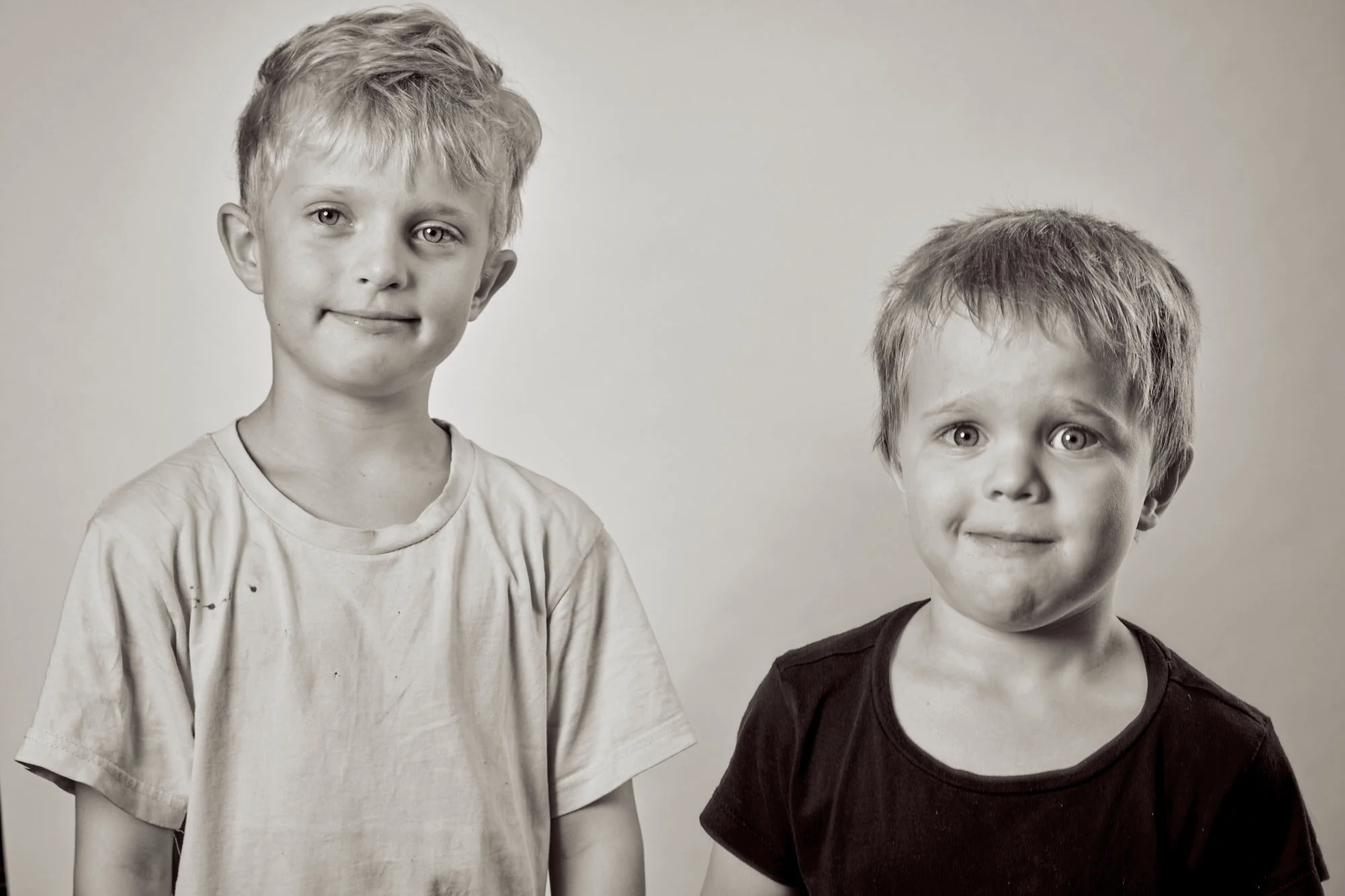 Black and white photo of two young boys with short, tousled hair, standing side by side against a plain background. The boy on the left is wearing a light-colored, slightly dirty t-shirt and has a subtle smile. The boy on the right is wearing a dark 