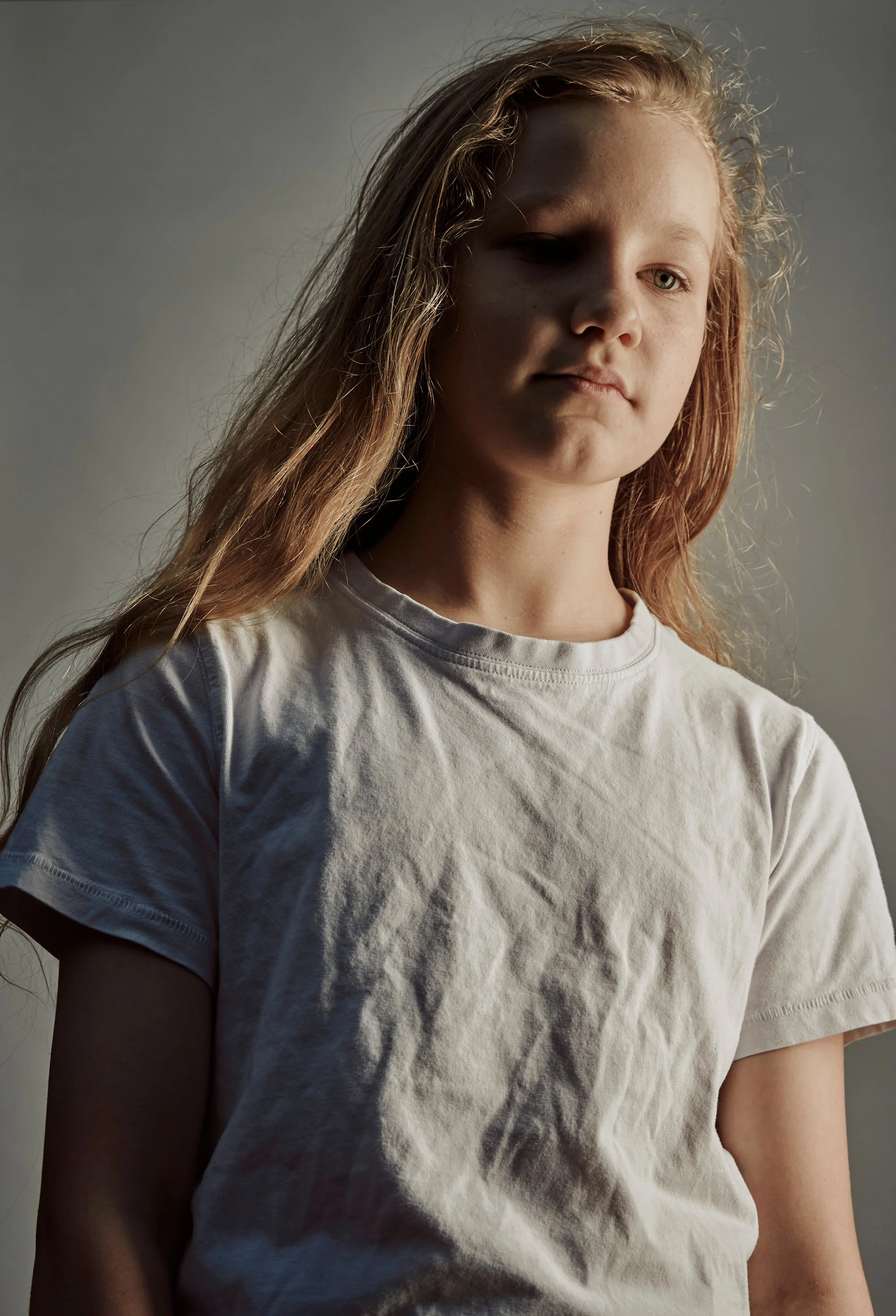 A teenage girl with long red hair wearing a white T-shirt, looking slightly down with a serious expression.