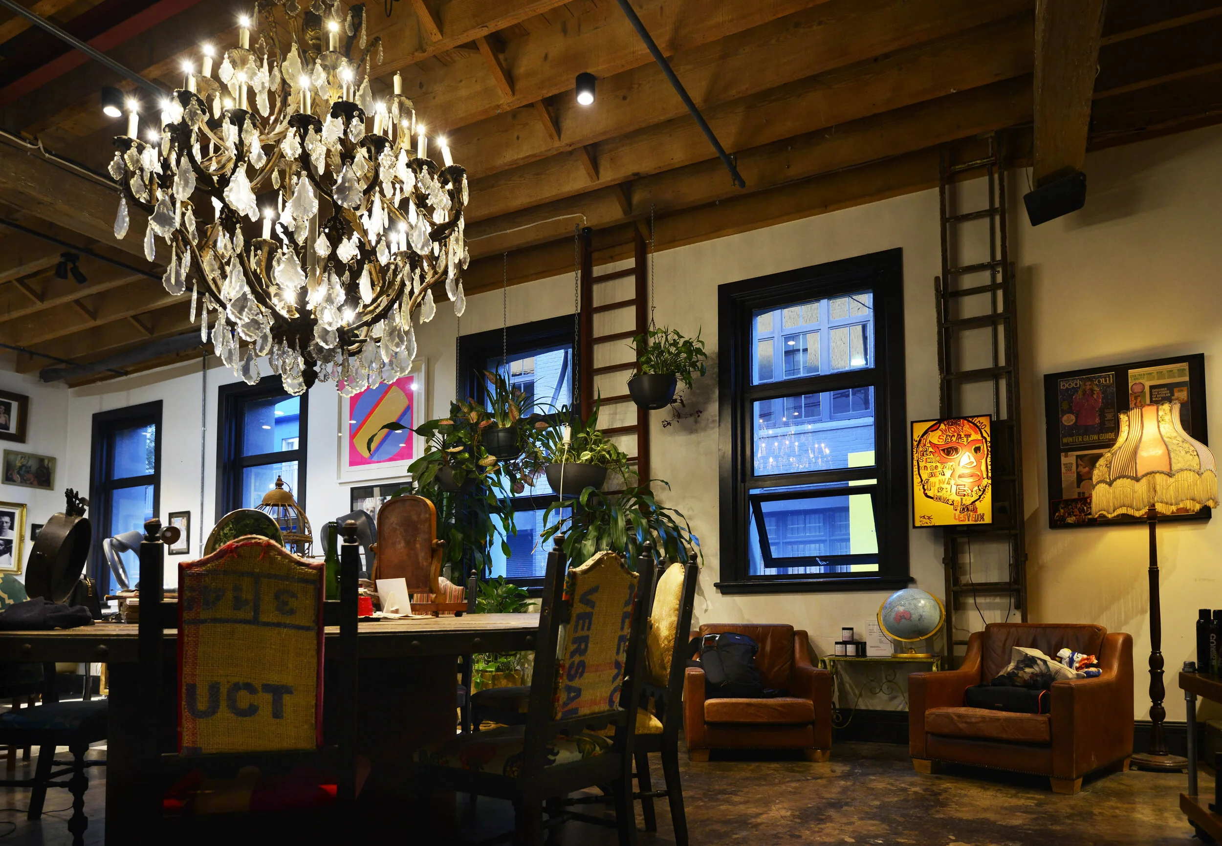 Interior of a cozy room with a wooden ceiling, a large chandelier, a table with chairs, armchairs, potted plants, window views of a cityscape, framed artwork, a vintage globe, and a standing lamp.