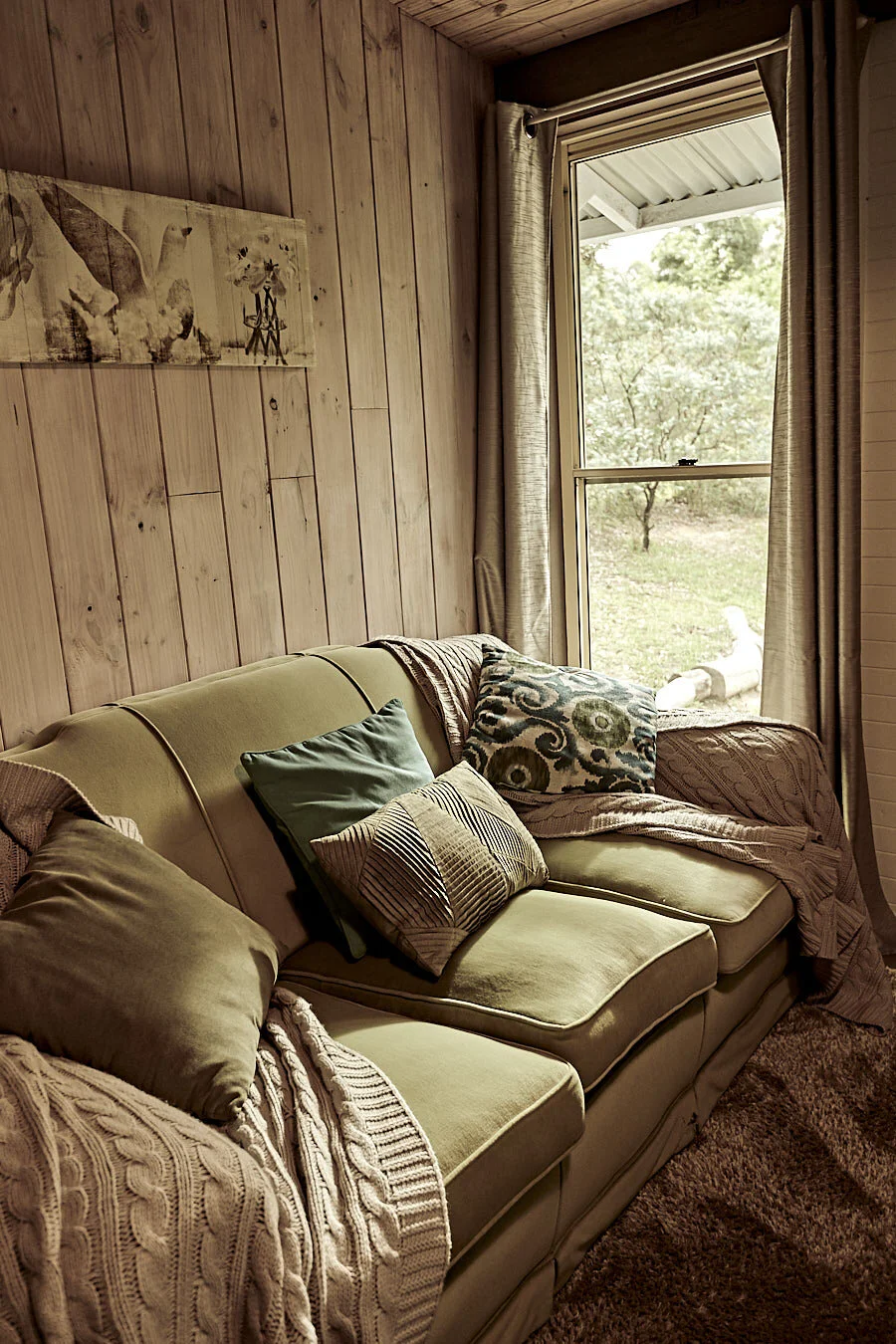 Living room with wooden wall, beige sofa with pillows and blankets, window with curtains showing trees outside.