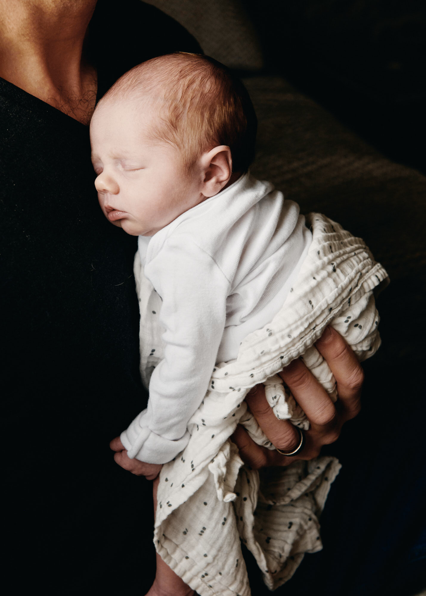A sleeping newborn baby wearing a white shirt and beige patterned pants, being held gently on their stomach by an adult with a dark shirt, against a dark background.