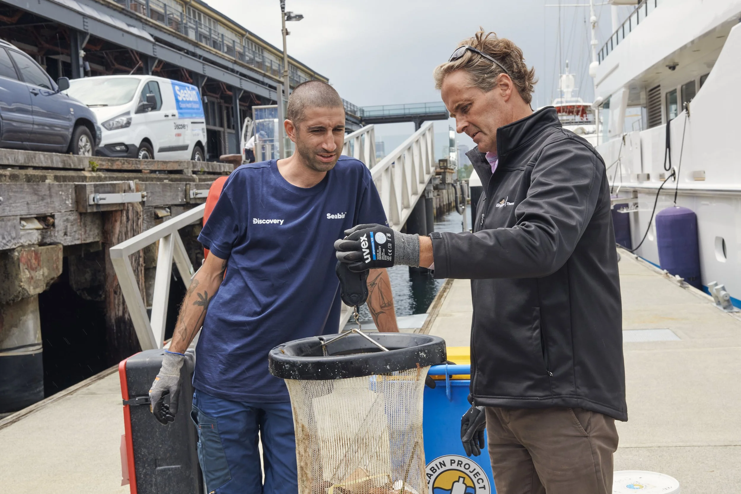 Two men working together on a dock, one showing a metal tool, a cleanup net, and a cooler. The man on the left is wearing a blue Discovery T-shirt and has tattoos and a shaved head. The man on the right is wearing a black jacket and gloves with a log