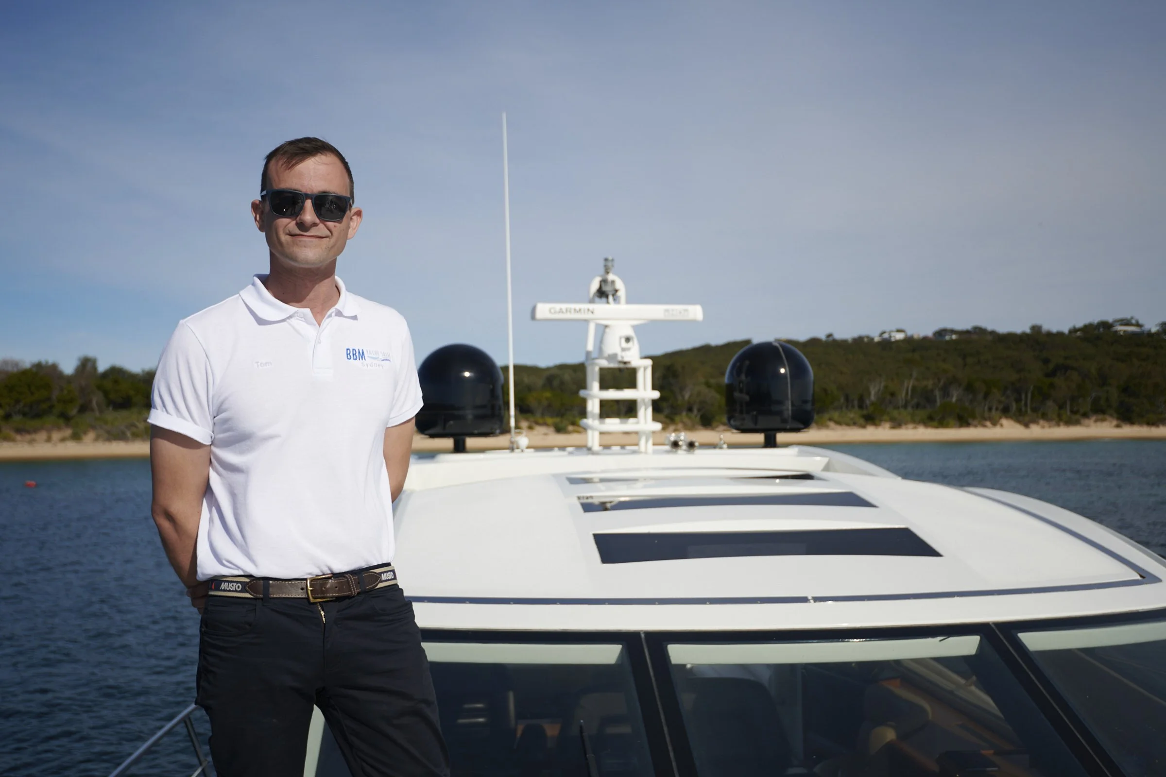 Man wearing sunglasses and a white polo shirt standing on a boat by the water with trees in the background.