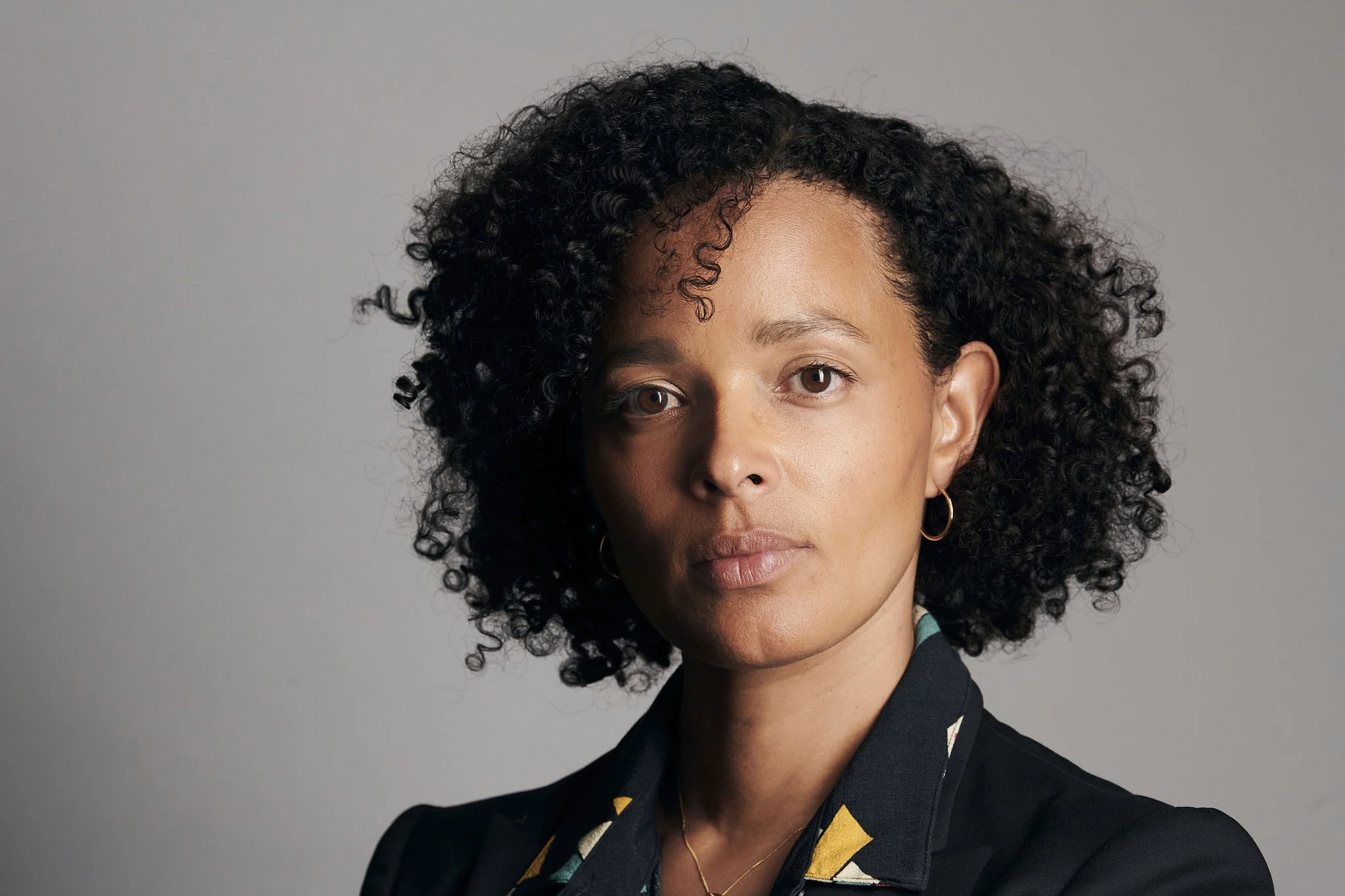 A woman with medium brown skin and curly black hair wearing a black blazer and gold hoop earrings, looking directly at the camera against a neutral gray background.