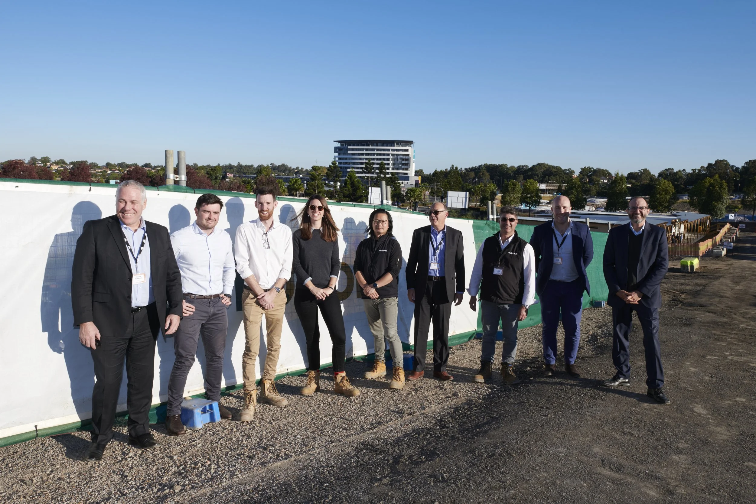 Group of ten people standing outdoors at a construction or development site, wearing business casual and casual attire, on a sunny day with a cityscape in the background.