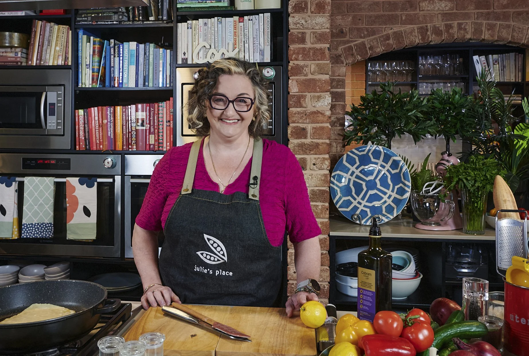 A woman with glasses and curly hair wearing a pink top and a black apron with the logo 'Julie's Place' standing in a kitchen with a variety of fresh vegetables, cooking utensils, and a pan on the stove.