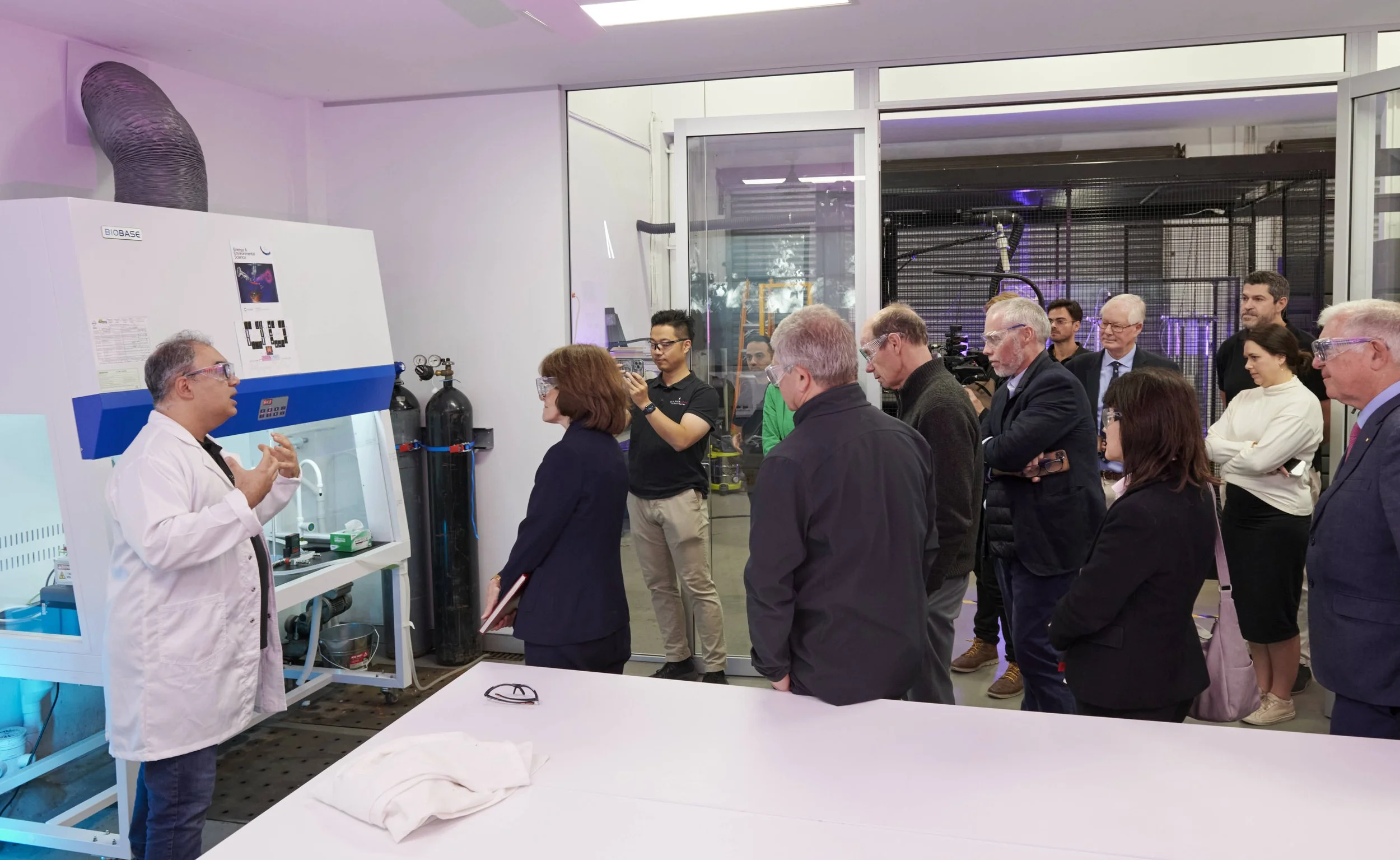 A man in a white lab coat and safety goggles is giving a presentation to a group of people inside a laboratory, with scientific equipment and large gas tanks in the background.