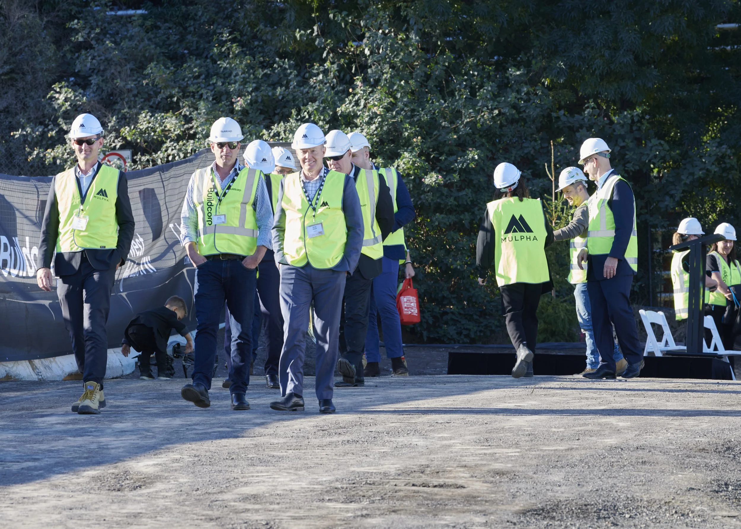 A group of people wearing safety helmets and vests walking on a construction site, with trees in the background.