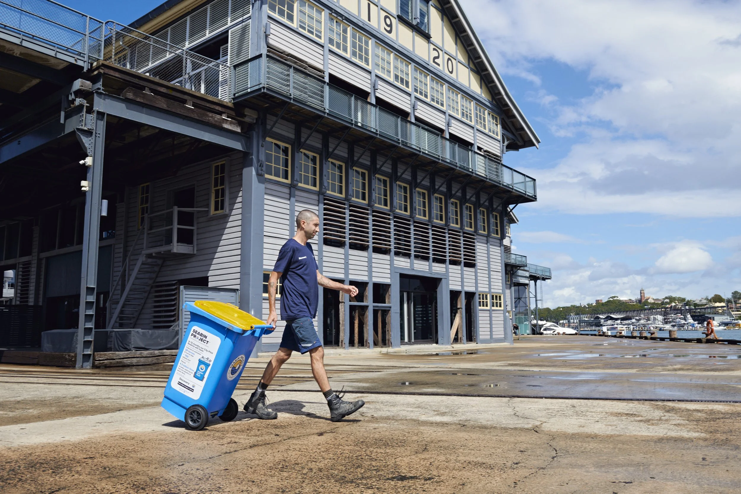 A man in a navy t-shirt and shorts with a tattoo on his left arm walks past a blue recycling bin, at a marina with a large industrial-style building and boats in the background.